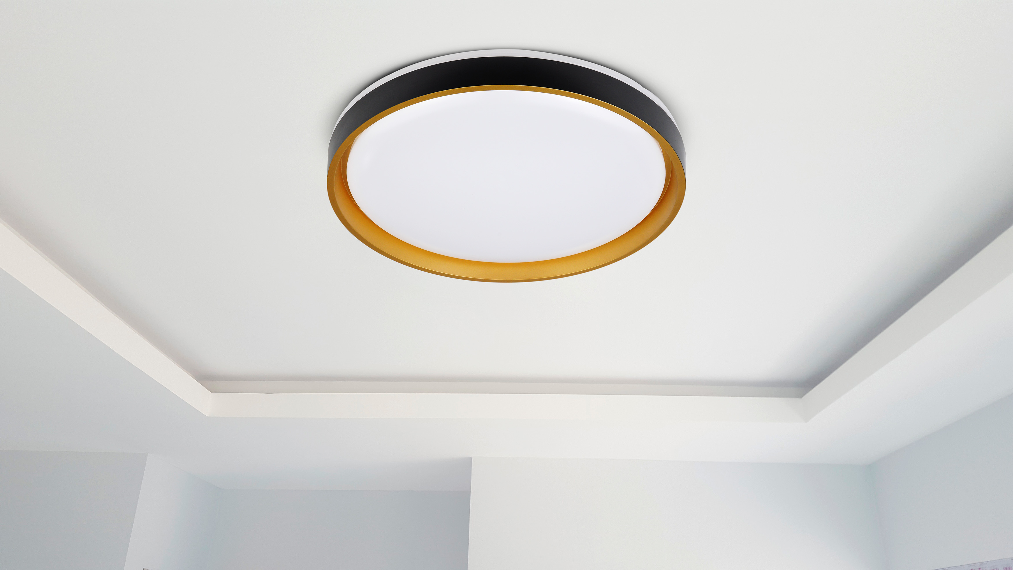 An einer weißen Decke ist eine runde Deckenleuchte montiert. Das Licht hat eine weiße Mitte, einen goldenen Ring und einen schwarzen Außenring.