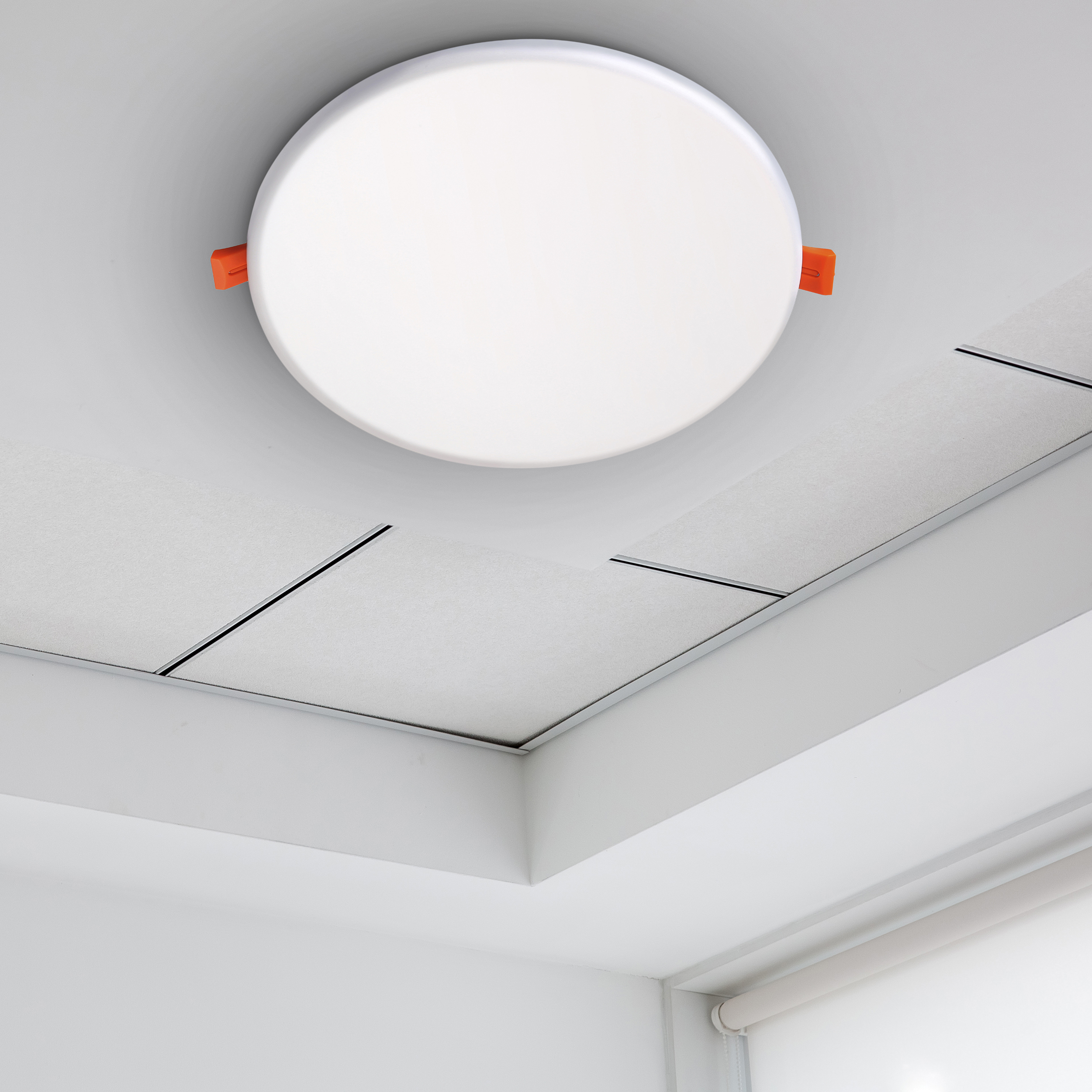 Eine runde, weiße Deckenleuchte mit orangefarbenen Clips ist an einer weißen Decke montiert. Weiße Deckenplatten und eine weiße Wand sind sichtbar.