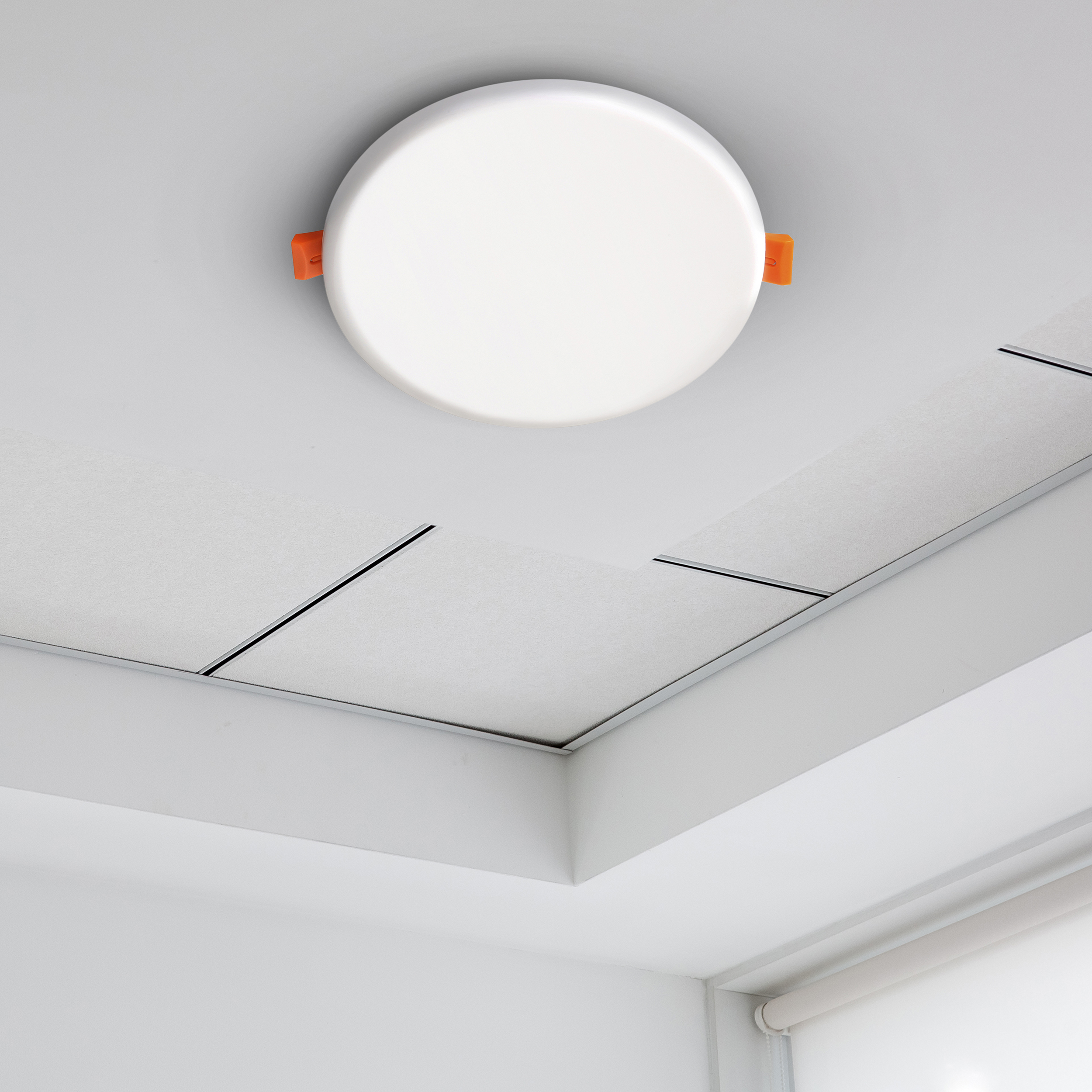 Eine kreisförmige, weiße Deckenleuchte mit orangefarbenen Clips ist an einer weißen Decke befestigt. Paneele und eine Ecke sind ebenfalls sichtbar.