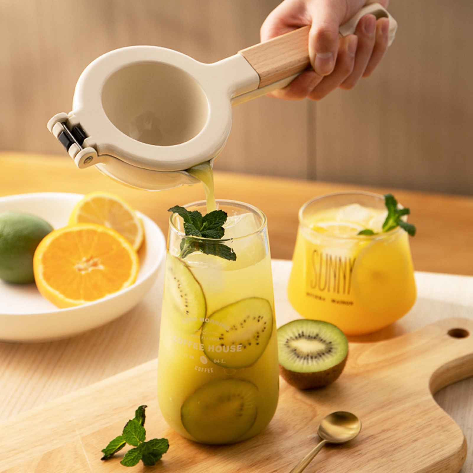 Eine Hand drückt eine Saftpresse und gießt Saft in ein Glas mit Kiwi und Minze. Andere Gläser und Früchte sind sichtbar.
