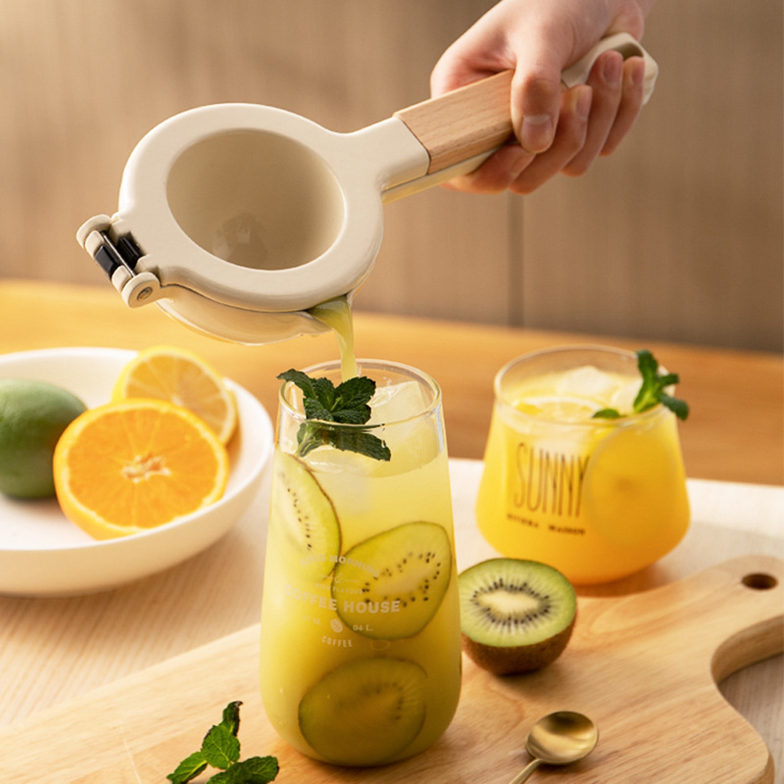 Eine Hand, die einen Entsafter über ein Glas Kiwisaft drückt. Scheiben von Kiwi und Orange, Minzblätter und ein Glas Orangensaft sind sichtbar.