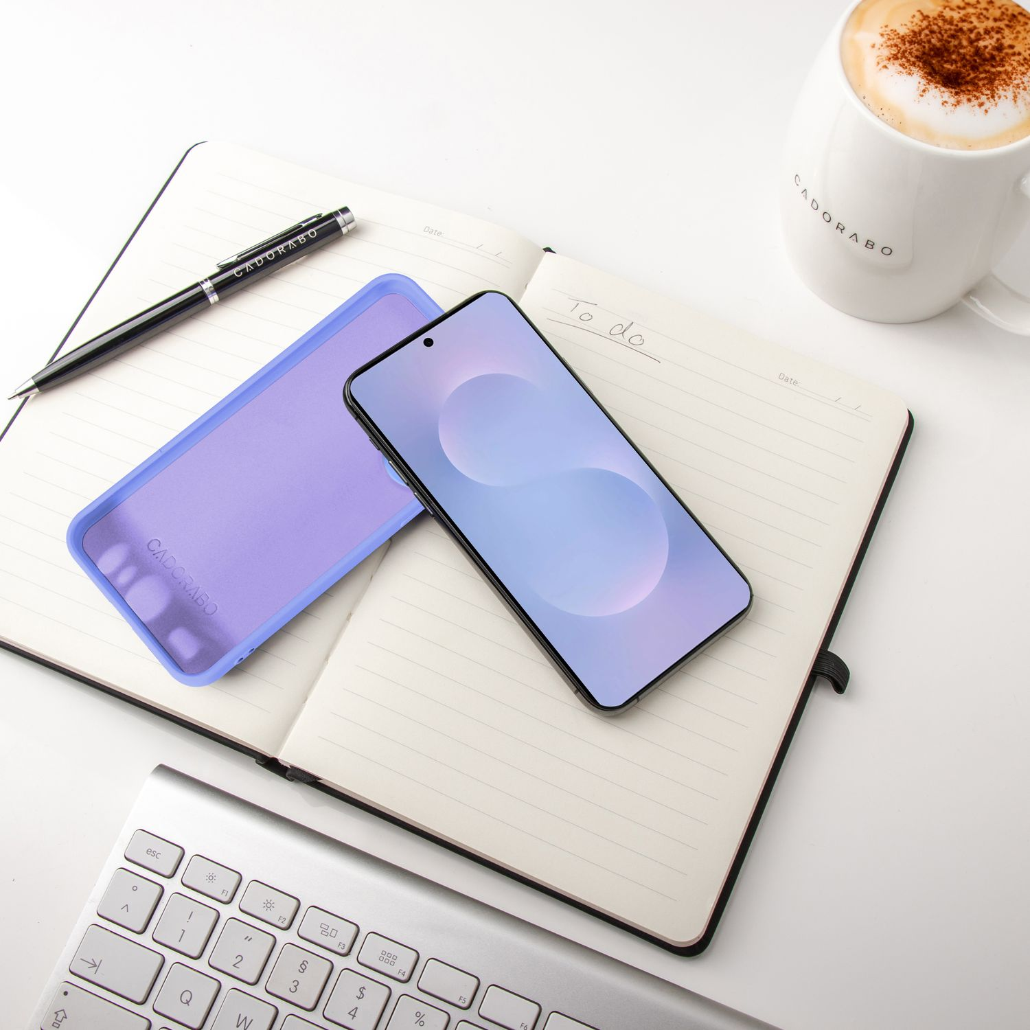 Ein Smartphone und eine Hülle auf einem offenen Notizbuch mit Stift und Kaffeetasse; Tastatur in der Ecke.