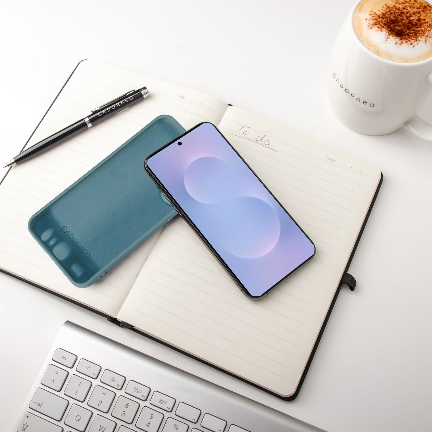 Smartphone, Hülle, Stift und Kaffeetasse auf offenem Notizbuch. Tastatur im Vordergrund. Weißer Hintergrund.