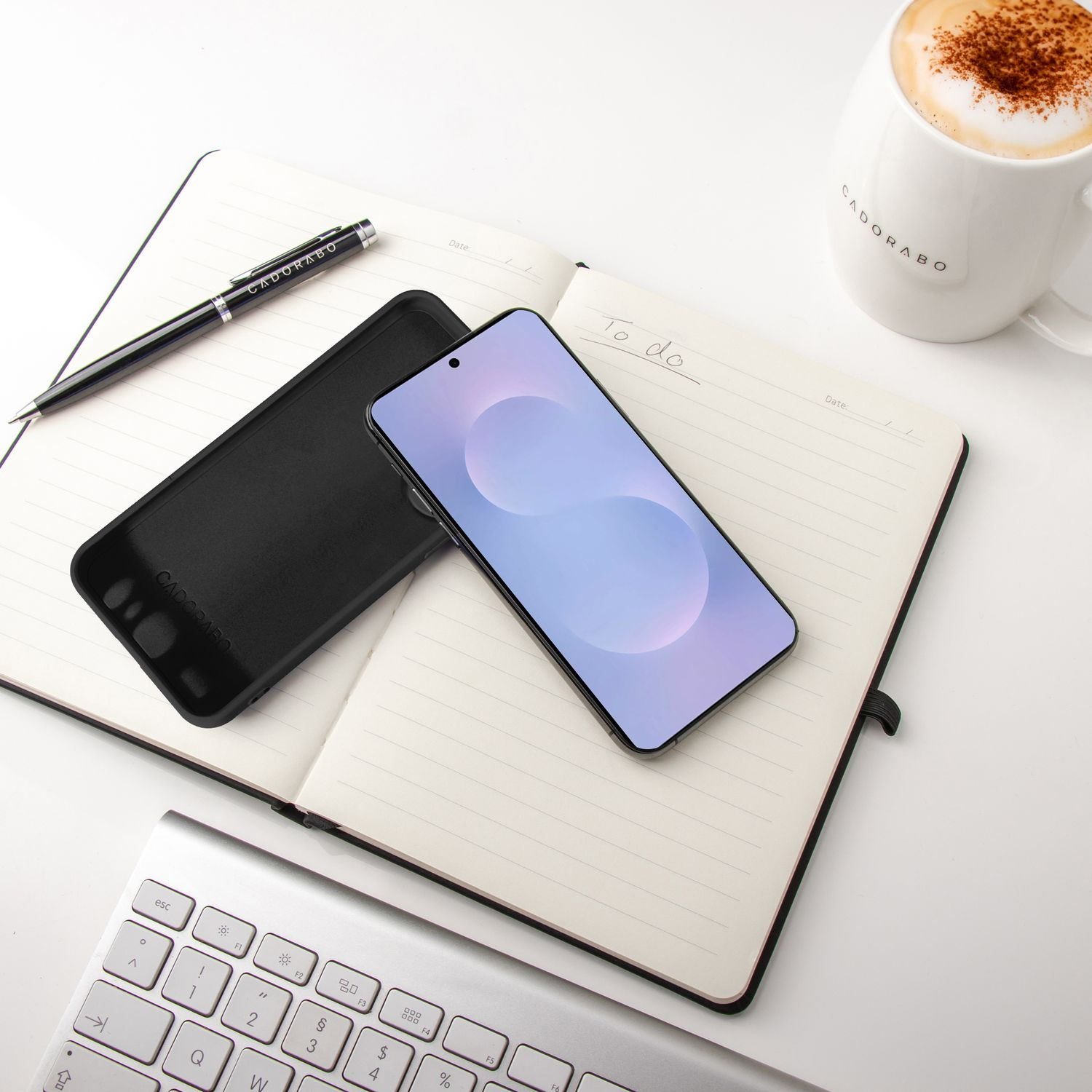 Ein Smartphone, eine Hülle, ein Stift und eine Kaffeetasse liegen auf einem Notizbuch. Eine Tastatur befindet sich im Vordergrund. Alles auf einer weißen Oberfläche.