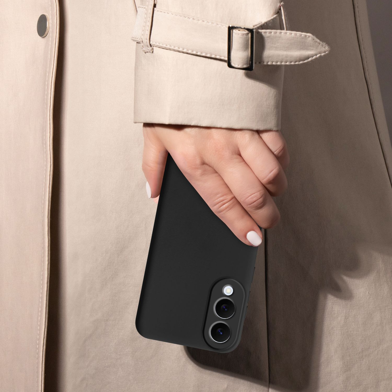 Eine Hand hält ein schwarzes Smartphone mit zwei Kameralinsen. Die Hand trägt weißen Nagellack und befindet sich neben einem beigefarbenen Mantel.
