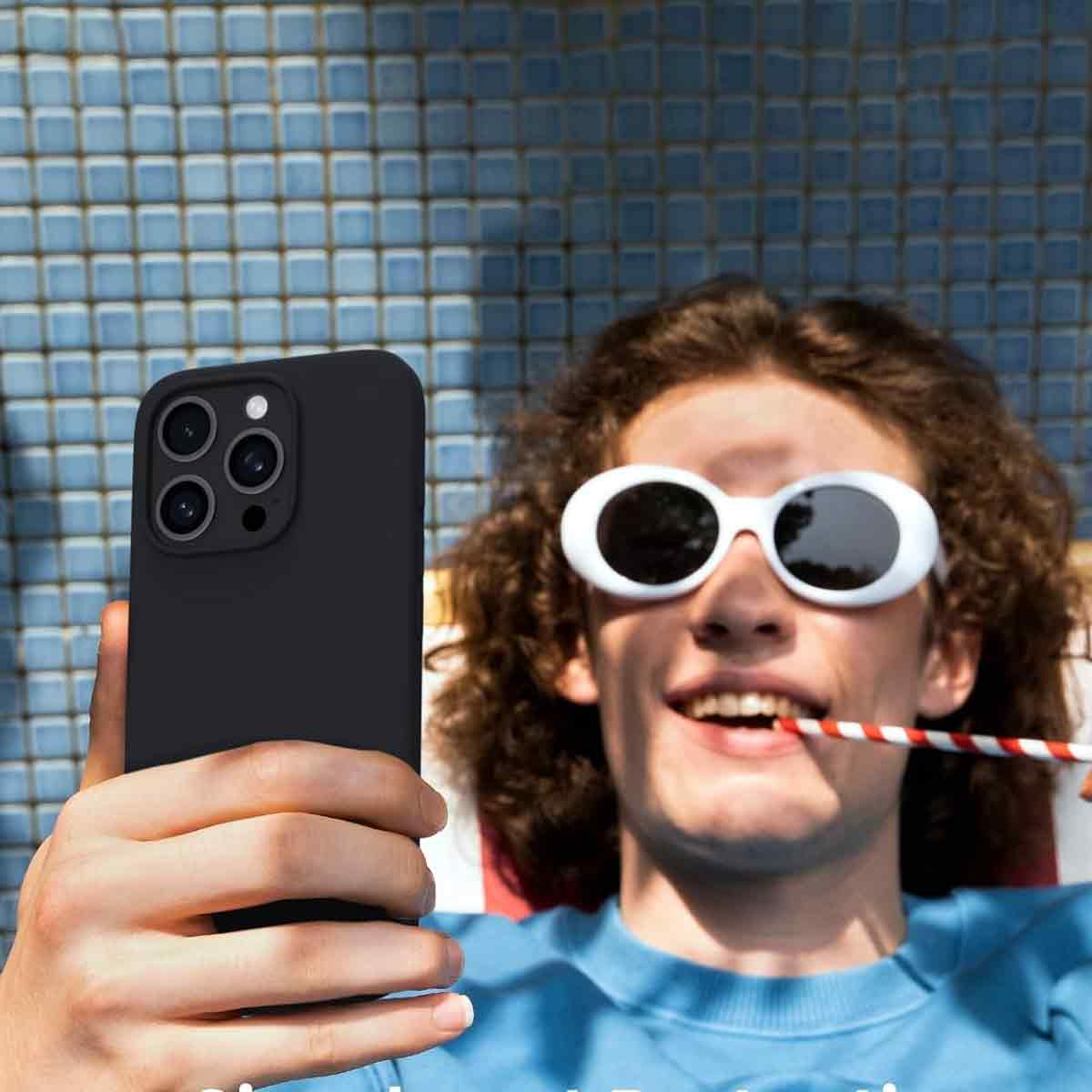 Eine Person mit Sonnenbrille hält ein Telefon. Sie lächelt mit einem Strohhalm im Mund. Der Hintergrund ist eine blaue Fliesenwand.