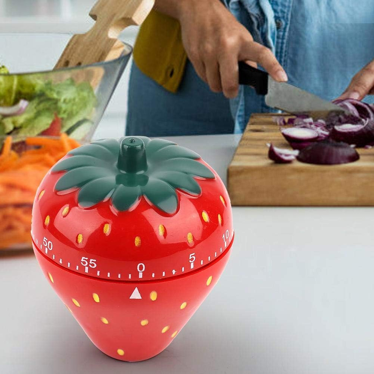 Ein roter Erdbeer-Timer steht auf einer Theke. Eine Person schneidet rote Zwiebeln auf einem Schneidebrett. Salat in einer Schüssel im Hintergrund.