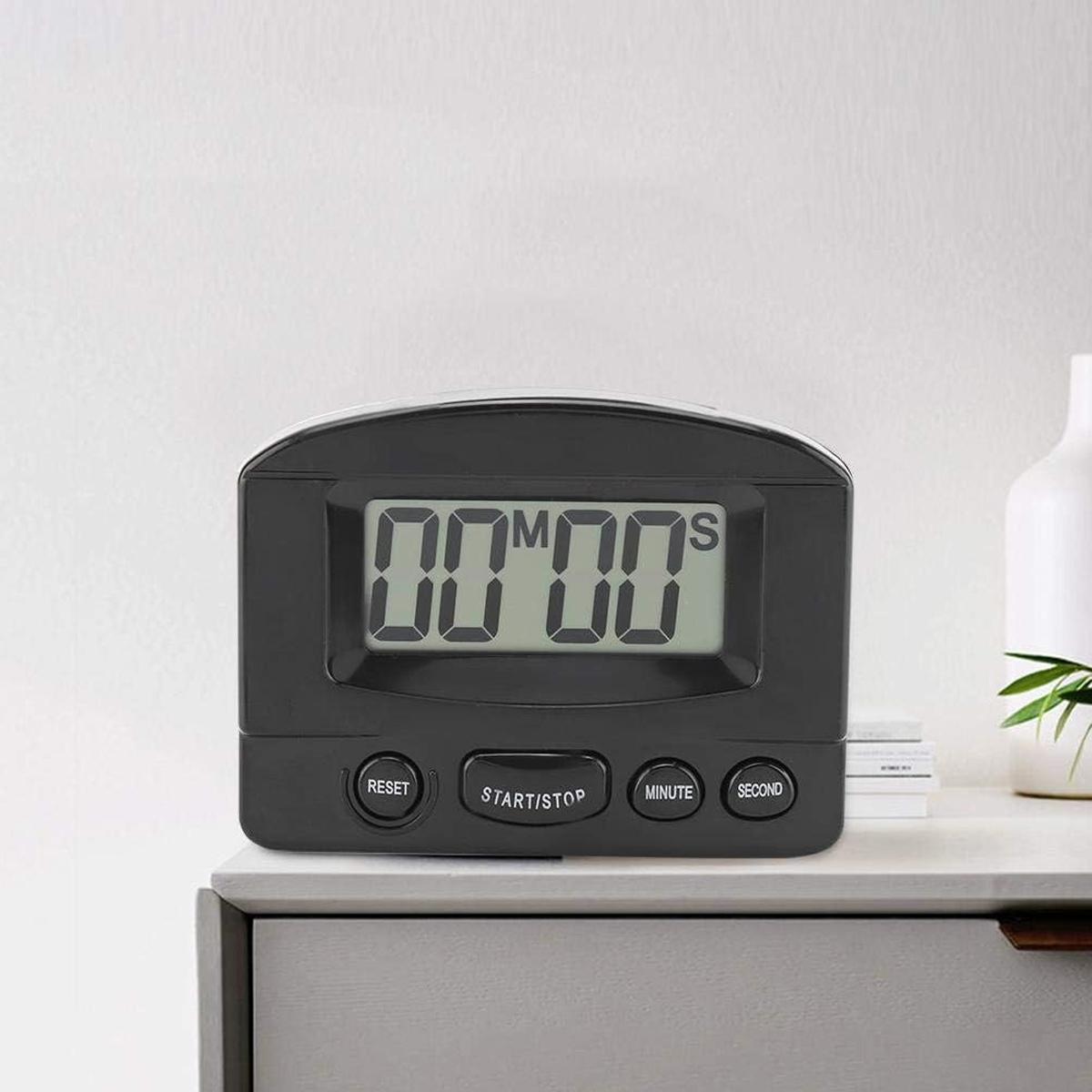 Ein schwarzer digitaler Küchentimer mit der Anzeige 00m00s liegt auf einer weißen Oberfläche, mit weißer Wand im Hintergrund und einer weißen Vase.