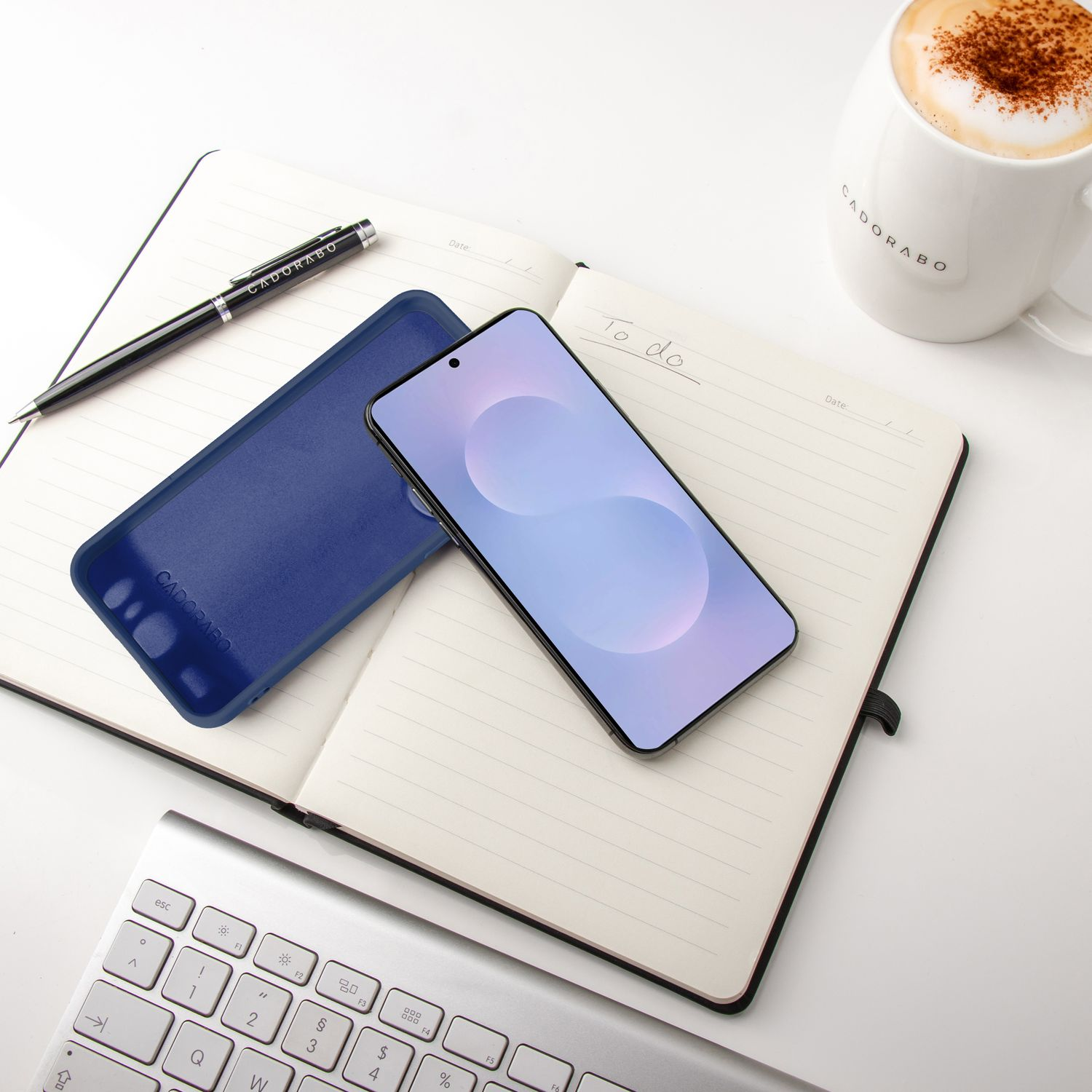 Ein Telefon auf einem offenen Notizbuch, ein Stift, eine blaue Handyhülle und eine Kaffeetasse auf einer weißen Oberfläche.