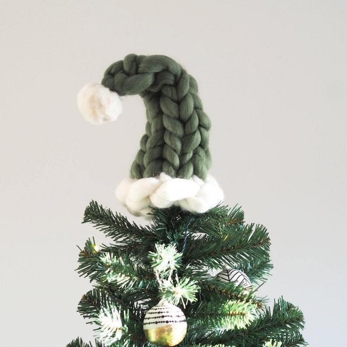 Ein Weihnachtsbaum mit einem grünen, gestrickten Hutaufsatz und Ornamenten. Der Hut ist olivgrün mit weißem Rand und Bommel.