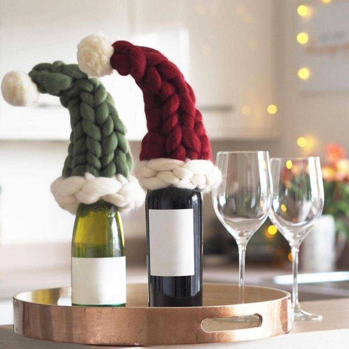 Zwei Weinflaschen mit Weihnachtsmützen auf einem Tablett, neben zwei Weingläsern. Eine Flasche ist grün, die andere rot. Der Hintergrund ist unscharf.