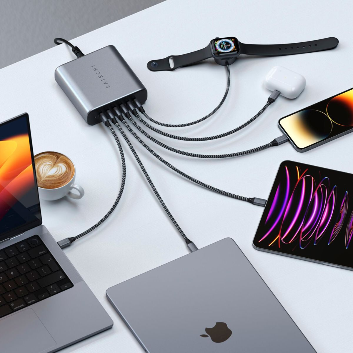 Un hub di ricarica argentato si collega a più dispositivi: laptop, tablet, telefono, orologio e AirPods. È presente anche una tazza di caffè.