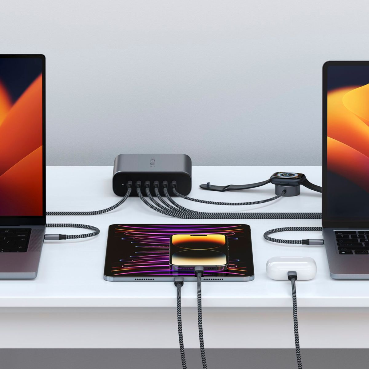 Stazione di ricarica con laptop, iPad, telefono, smartwatch e AirPods. Cavi grigi collegano i dispositivi a un hub grigio su una superficie bianca.