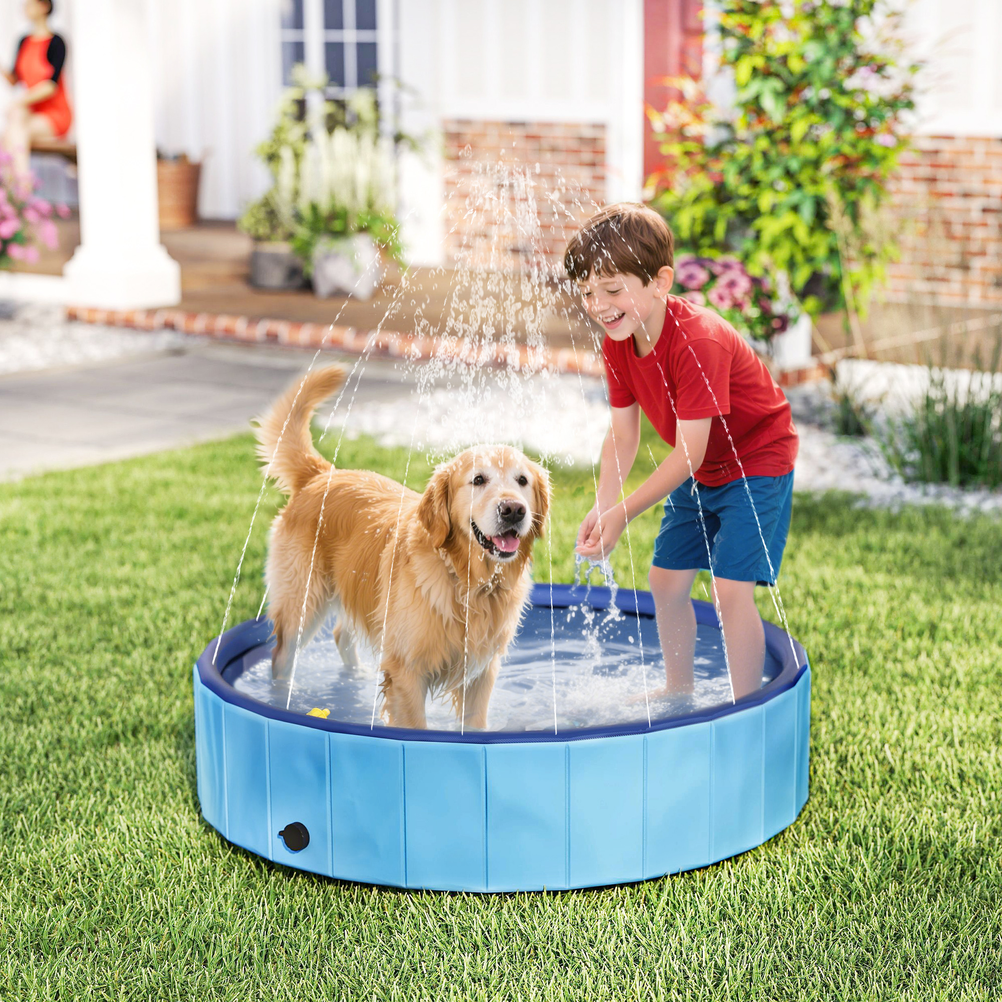Un golden retriever y un niño juegan en una piscina azul. El agua sale de arriba. Una mujer está sentada en el porche.