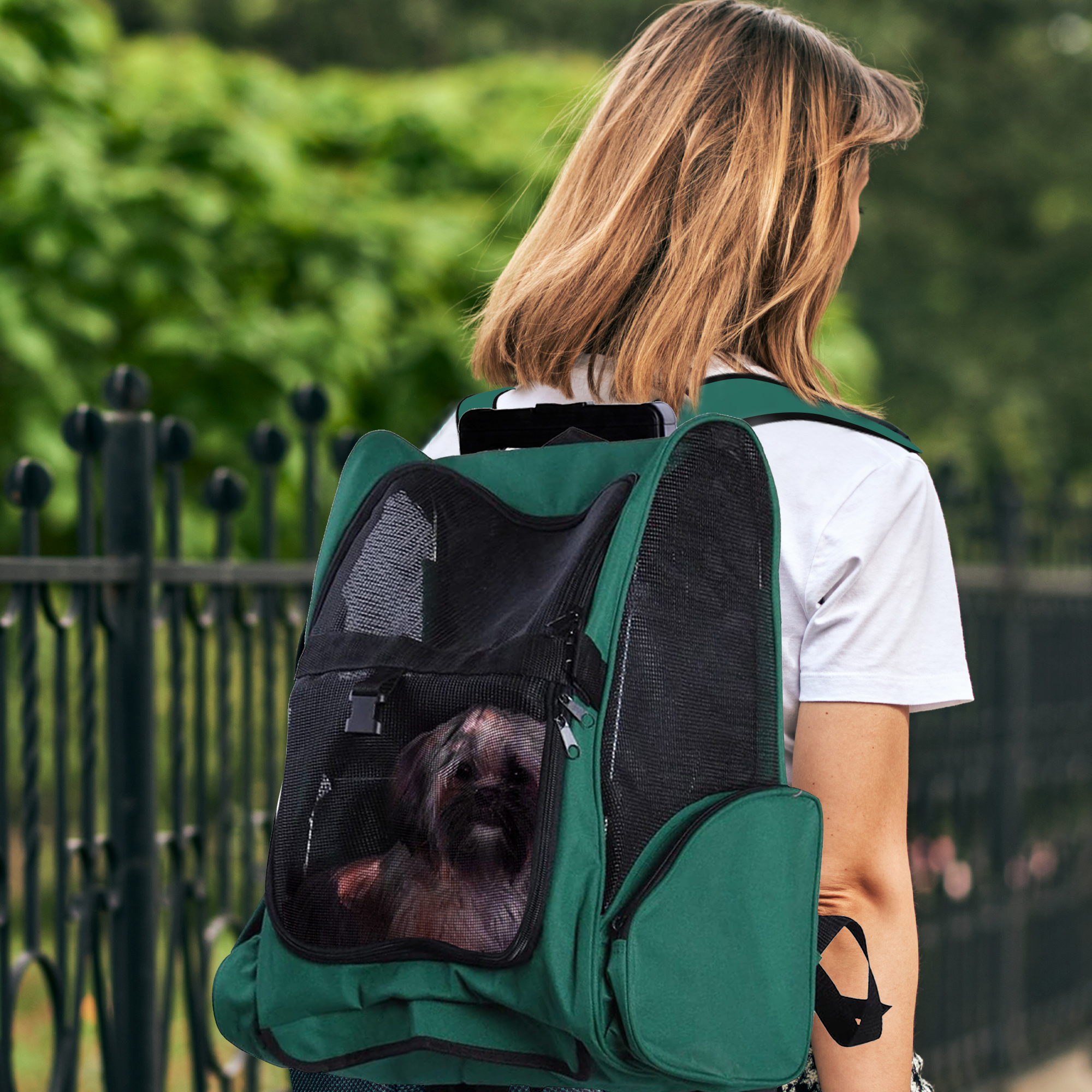 Mujer con cabello rubio, camisa blanca, lleva una mochila verde con un perro dentro. Valla negra y árboles verdes al fondo.