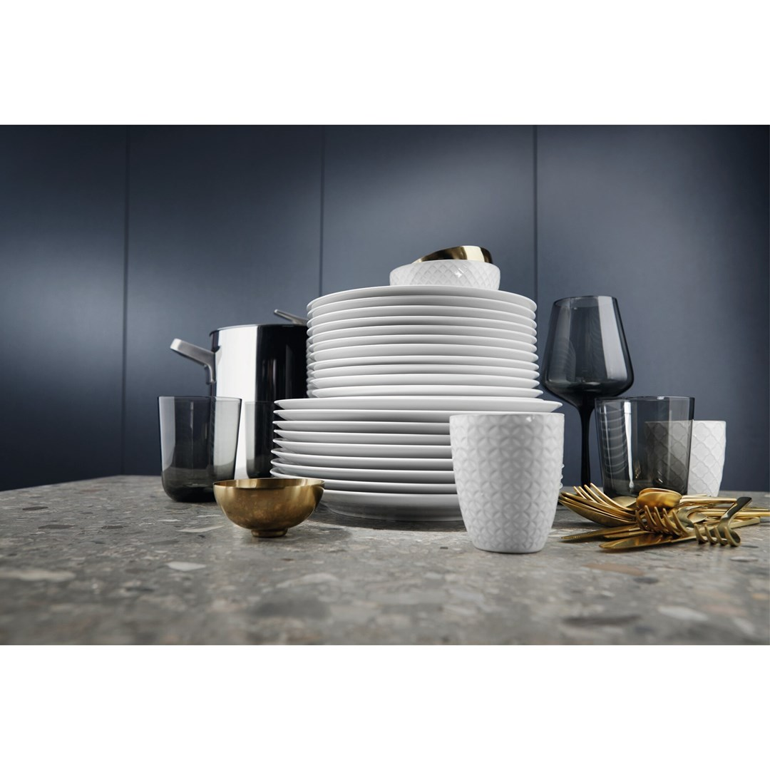 Nakrycie stołu z białymi talerzami, szklankami, złotymi sztućcami i garnkiem na nakrapianym blacie. Ciemnoniebieskie tło.