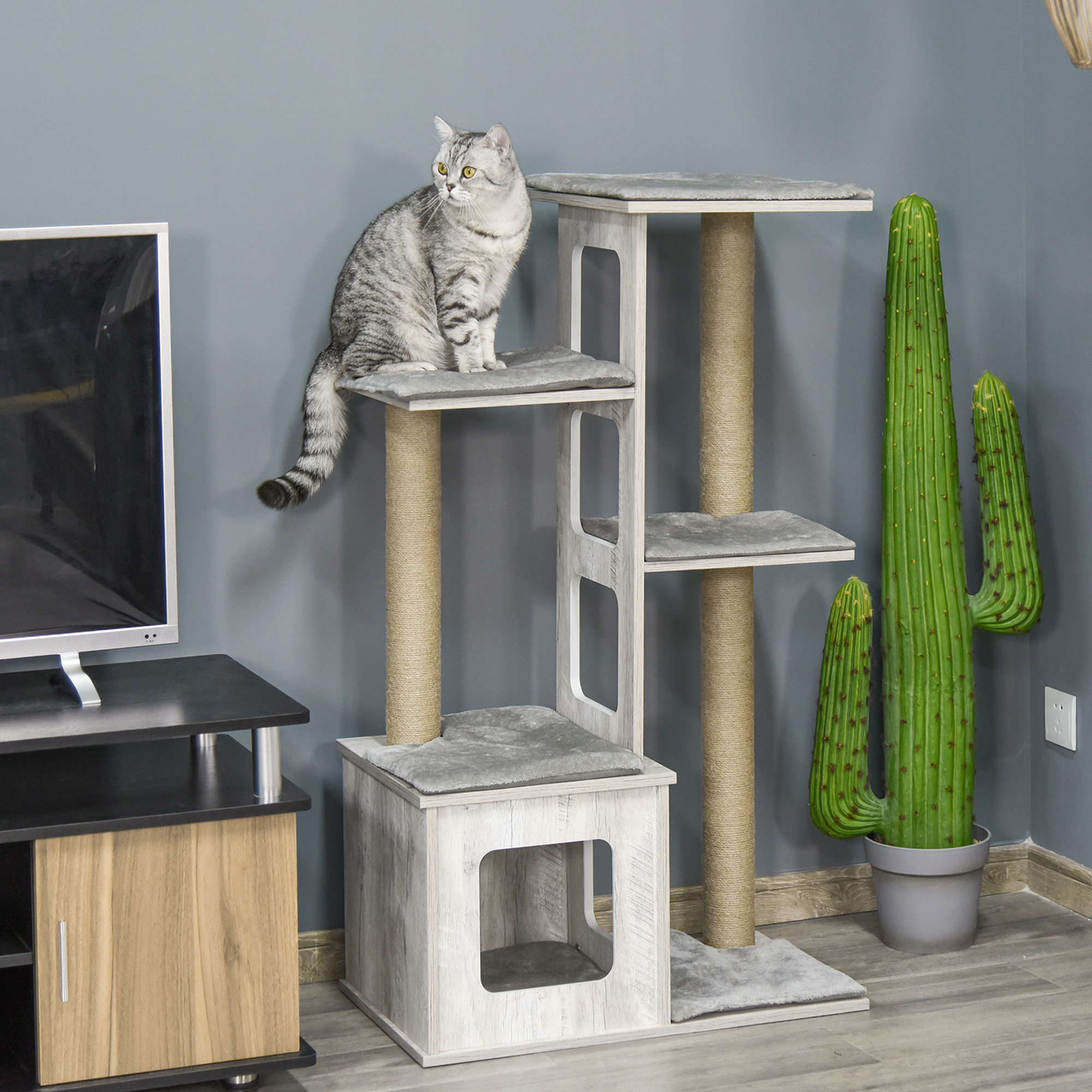 Un gato está sentado en un rascador. Un televisor y un cactus también están en la habitación. El gato es gris. El rascador es de madera.