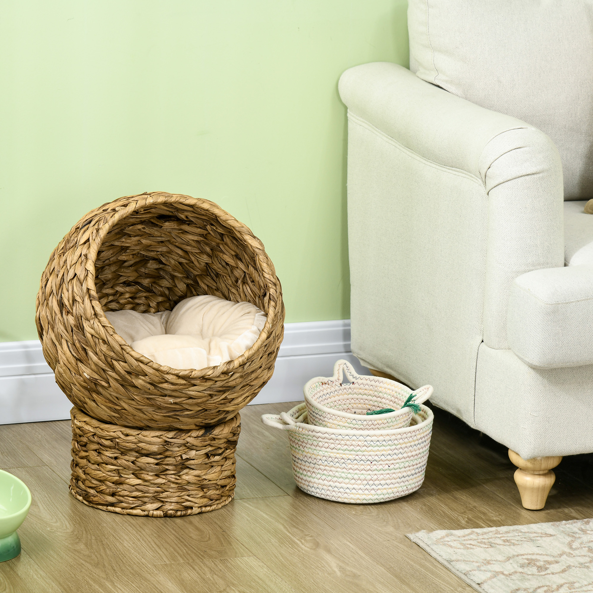 Una cama redonda tejida para gatos, dos cestas pequeñas y un sofá están sobre un piso de madera. La pared es verde claro.