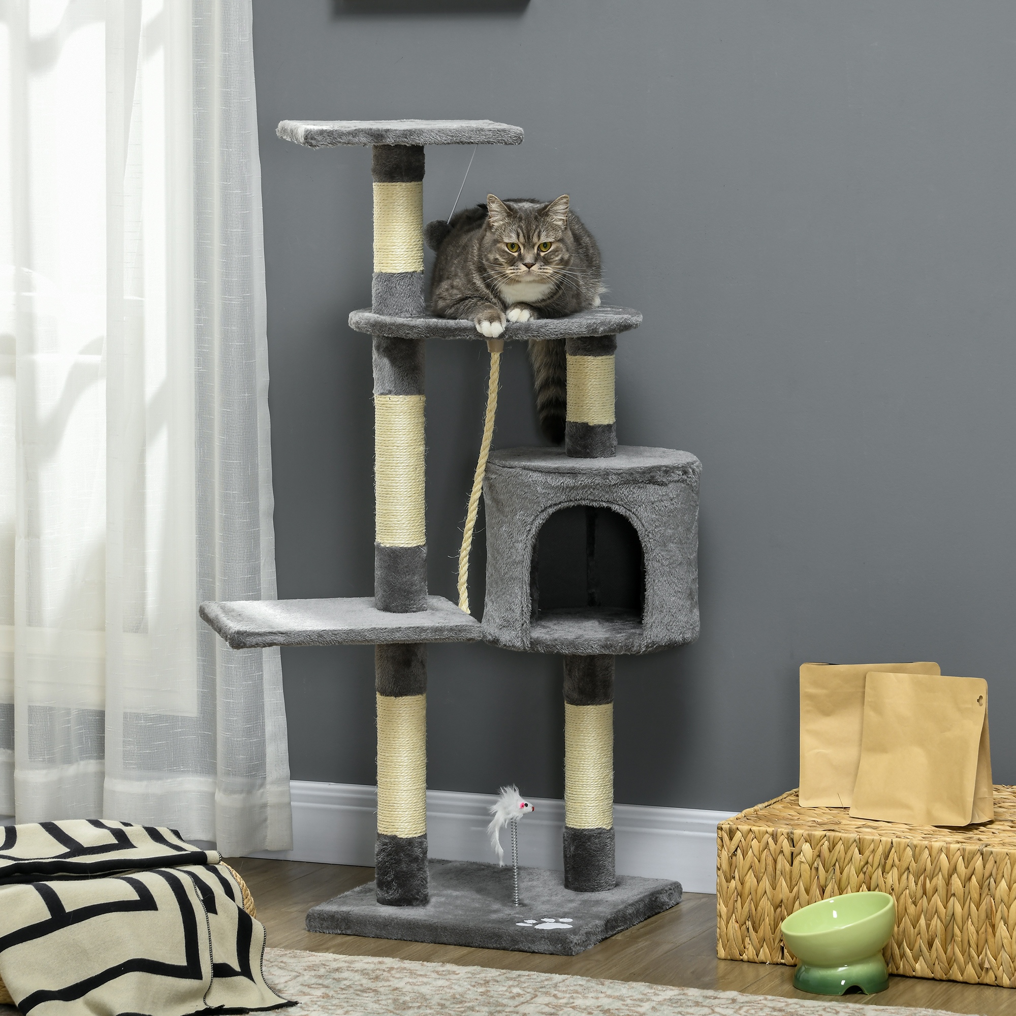 Un gato está sentado en un árbol para gatos gris. Manta, cesta y cuenco de comida en primer plano. Ventana y pared de fondo.