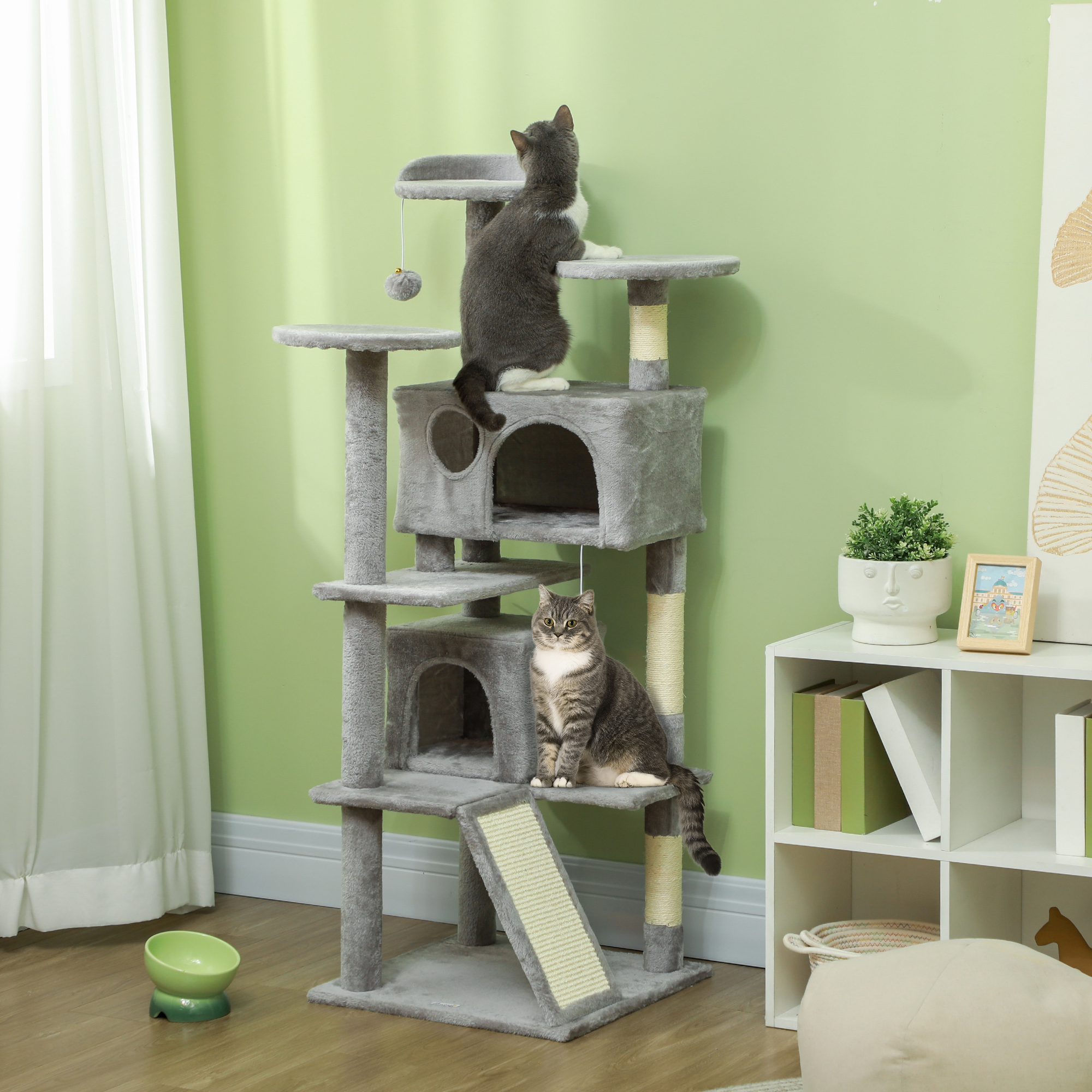 Dos gatos en un árbol para gatos de varios niveles. El árbol es gris, contra una pared verde. Un estante blanco tiene decoraciones.