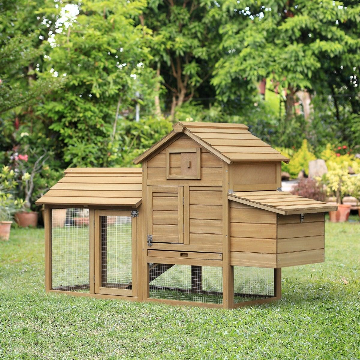 Un gallinero de madera con un recinto de malla metálica se encuentra sobre la hierba, rodeado de árboles y macetas.