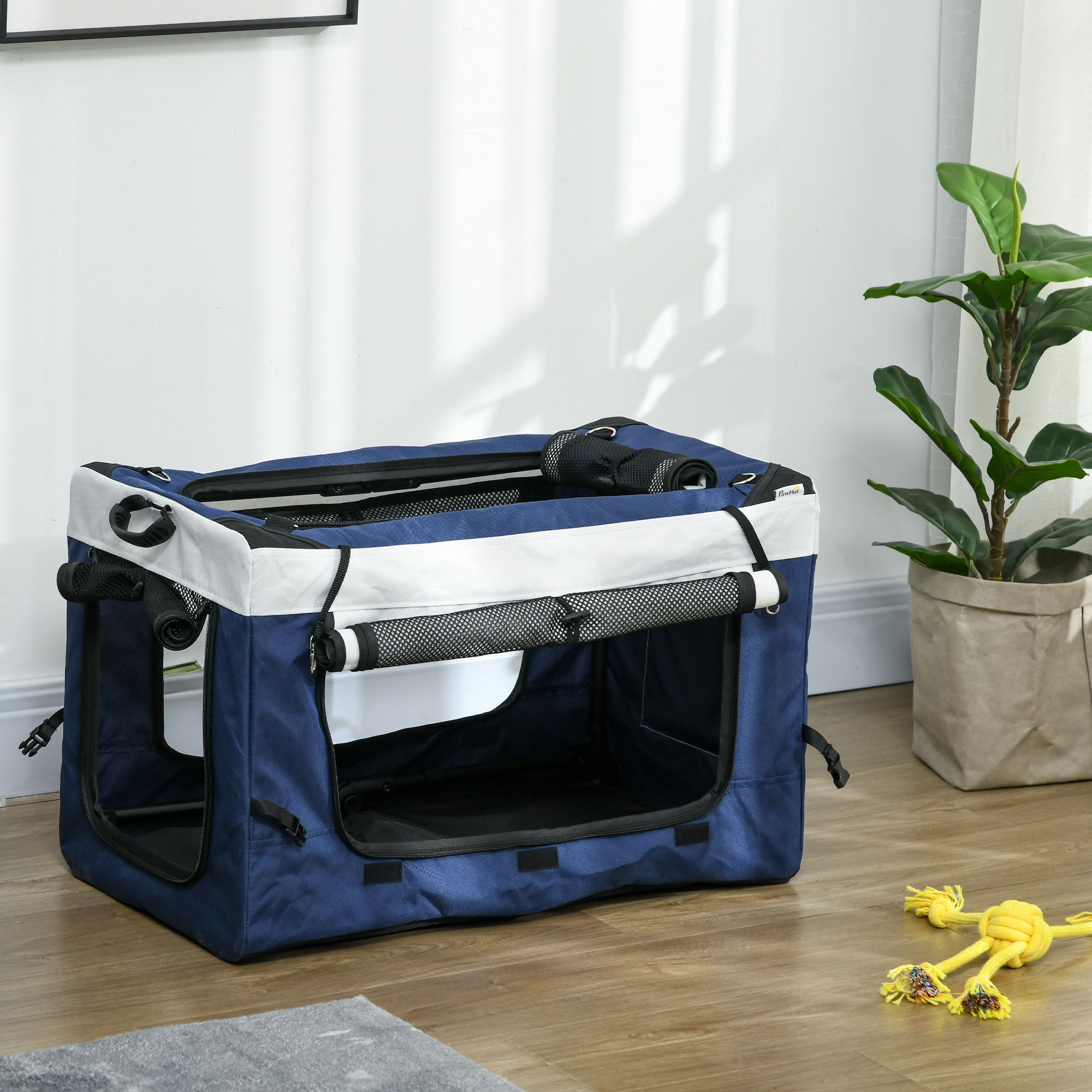 Una jaula azul y blanca para perros está en un piso de madera. Un juguete amarillo para perros está cerca, con una planta en el fondo.