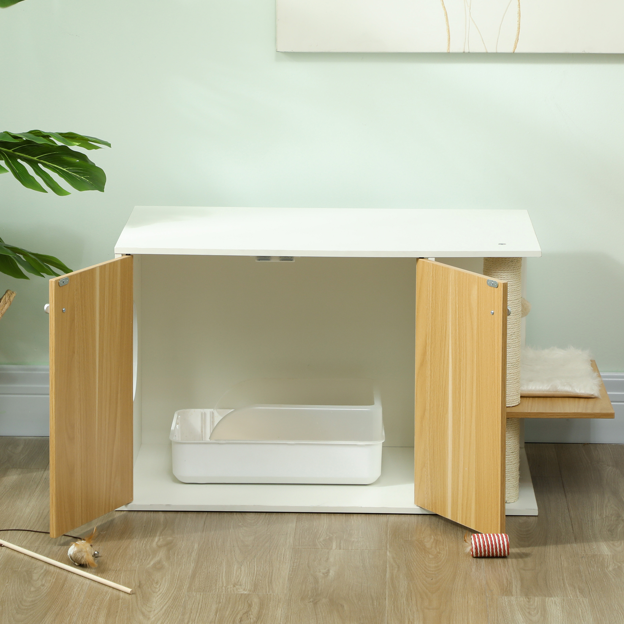 Un arenero blanco con puertas de madera. Un rascador para gatos y un juguete en el suelo. Una planta a la izquierda.