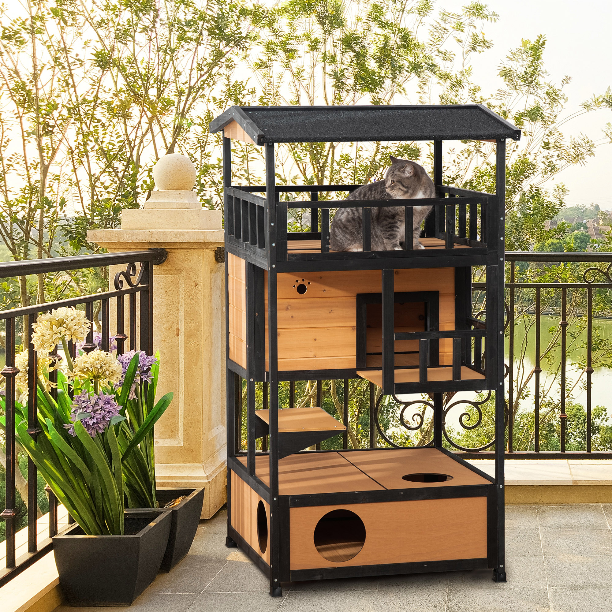 Una casa para gatos de varios niveles en un patio. Un gato gris está en el nivel superior. Plantas y un lago al fondo.