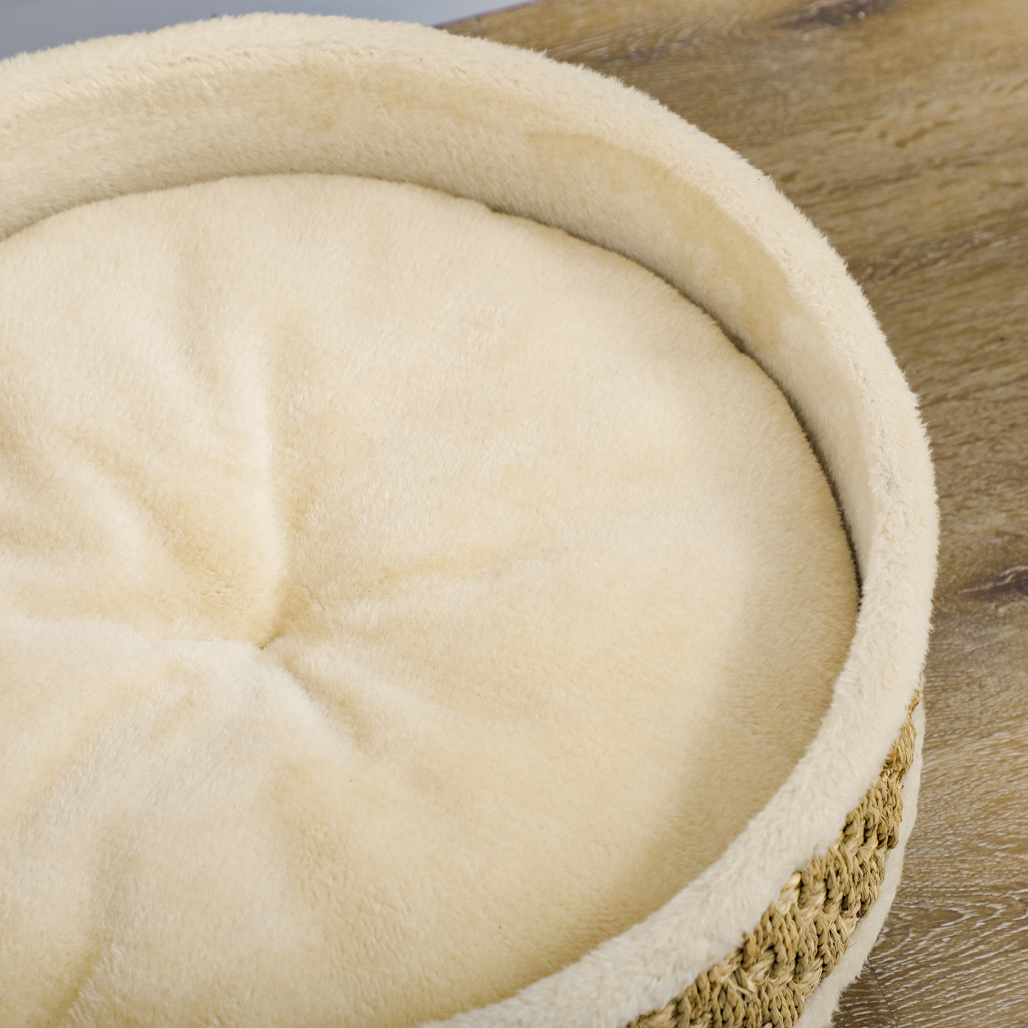 Primer plano de una cama beige para mascotas. Tiene un interior suave y esponjoso y un exterior tejido de color canela. La cama está sobre una superficie con vetas de madera.
