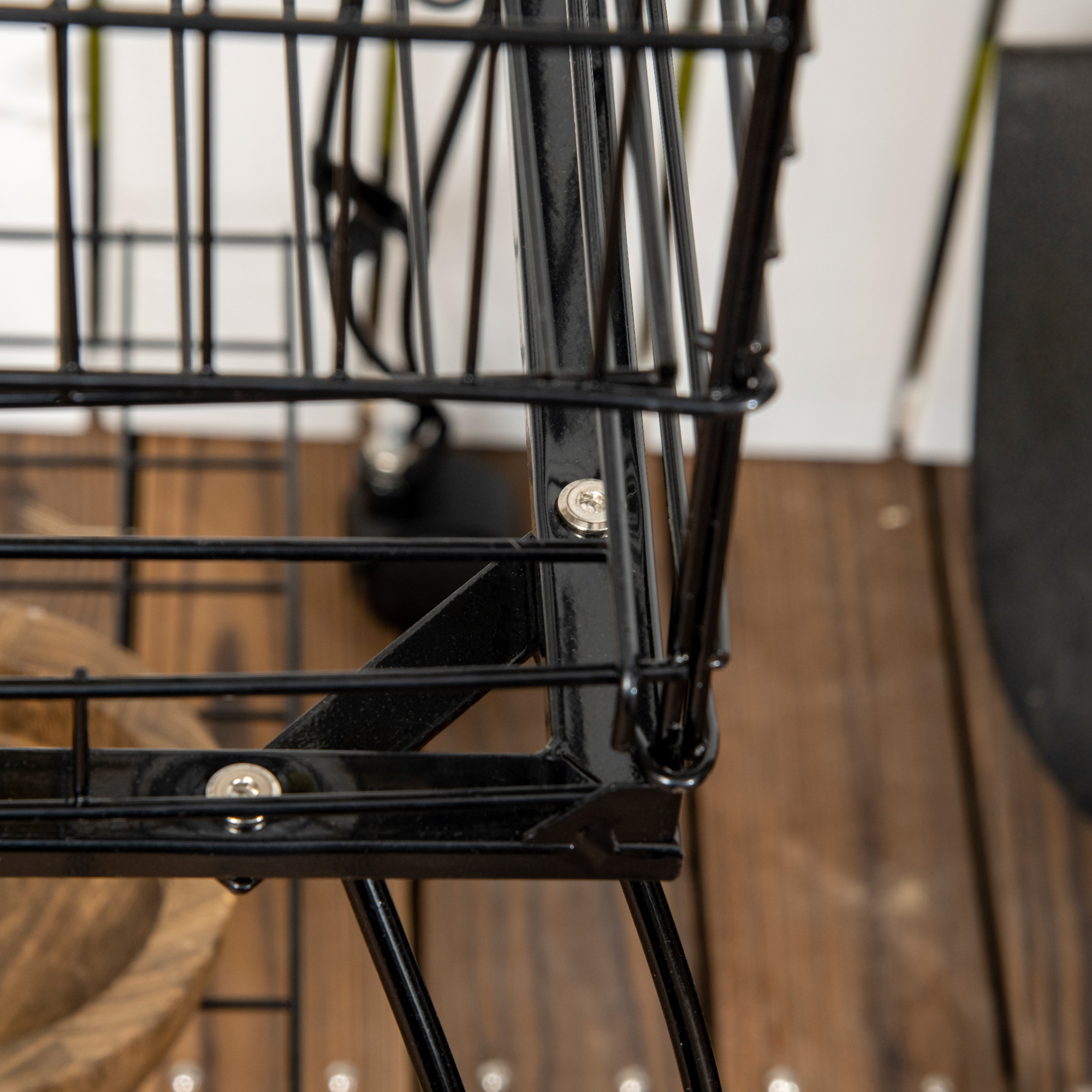 Primer plano de un carrito de compras de alambre negro. Piso de madera en el fondo. Fijaciones metálicas visibles, detalles de la estructura del carrito.