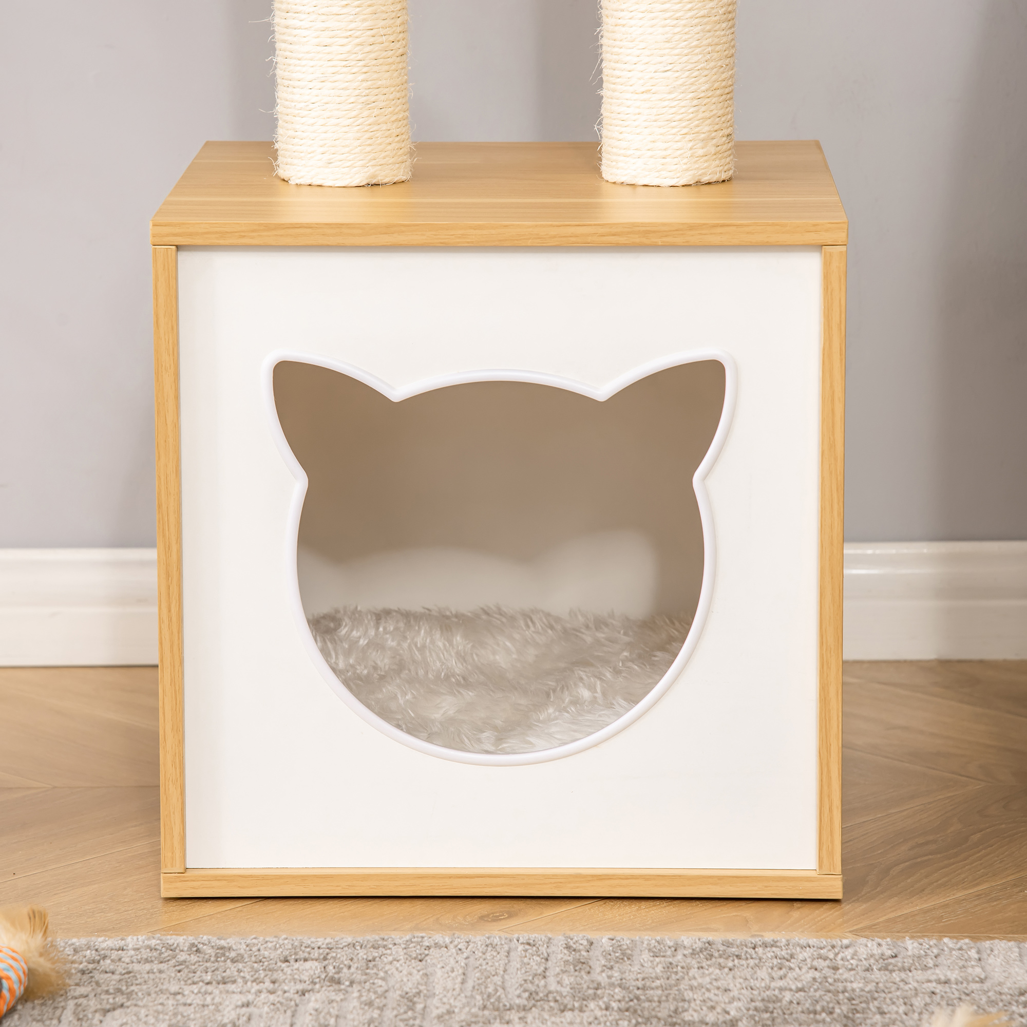 Casa para gatos con abertura en forma de gato. Tiene dos postes rascadores arriba. Casa blanca con marco de madera y cama peluda.