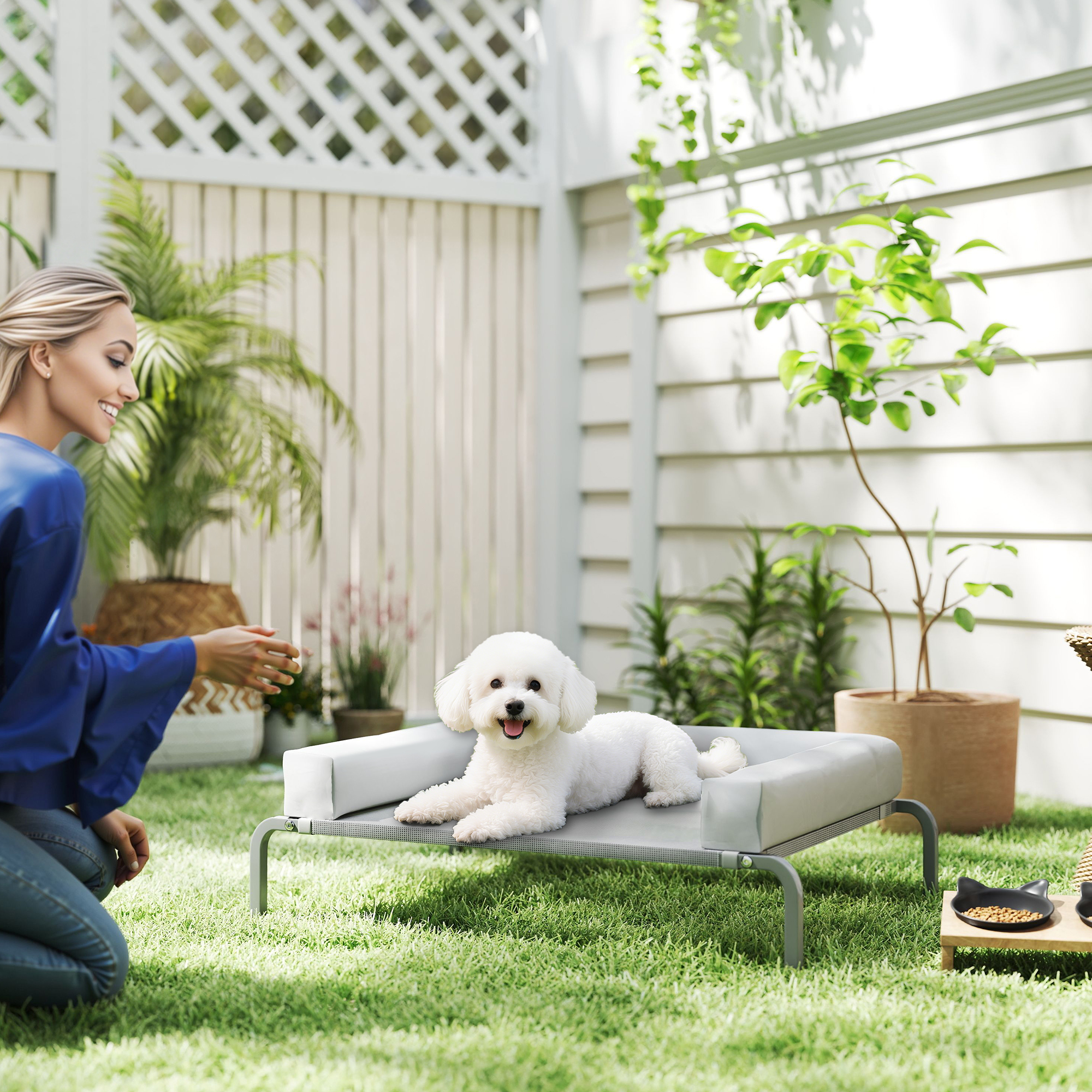 Una mujer se arrodilla, extendiendo la mano hacia un perro blanco en una cama en un jardín. Se ven hierba verde, plantas y una valla blanca.