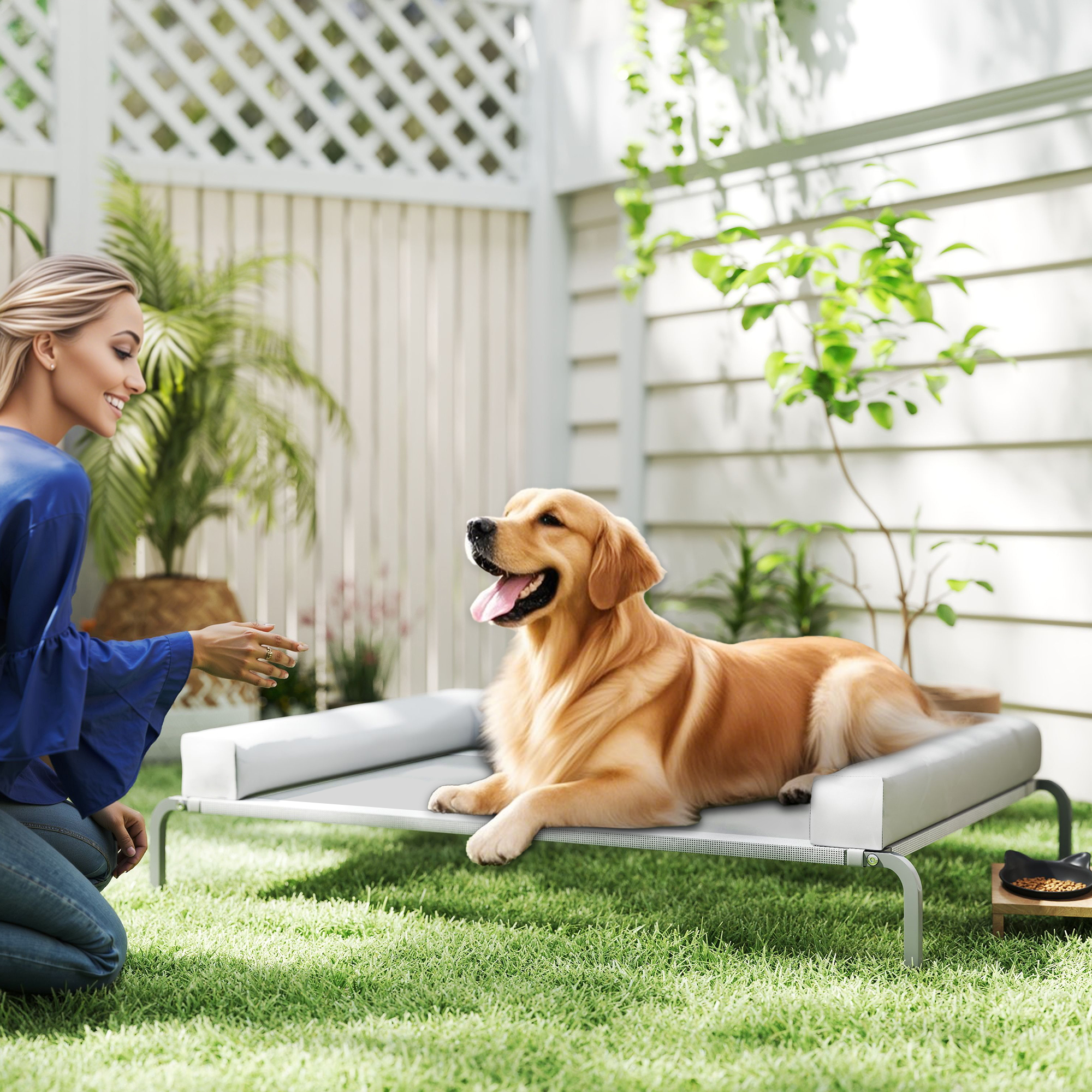 Mujer de azul con un golden retriever en una cama al aire libre. Césped y valla blanca en el fondo. Cuenco para perros cerca.
