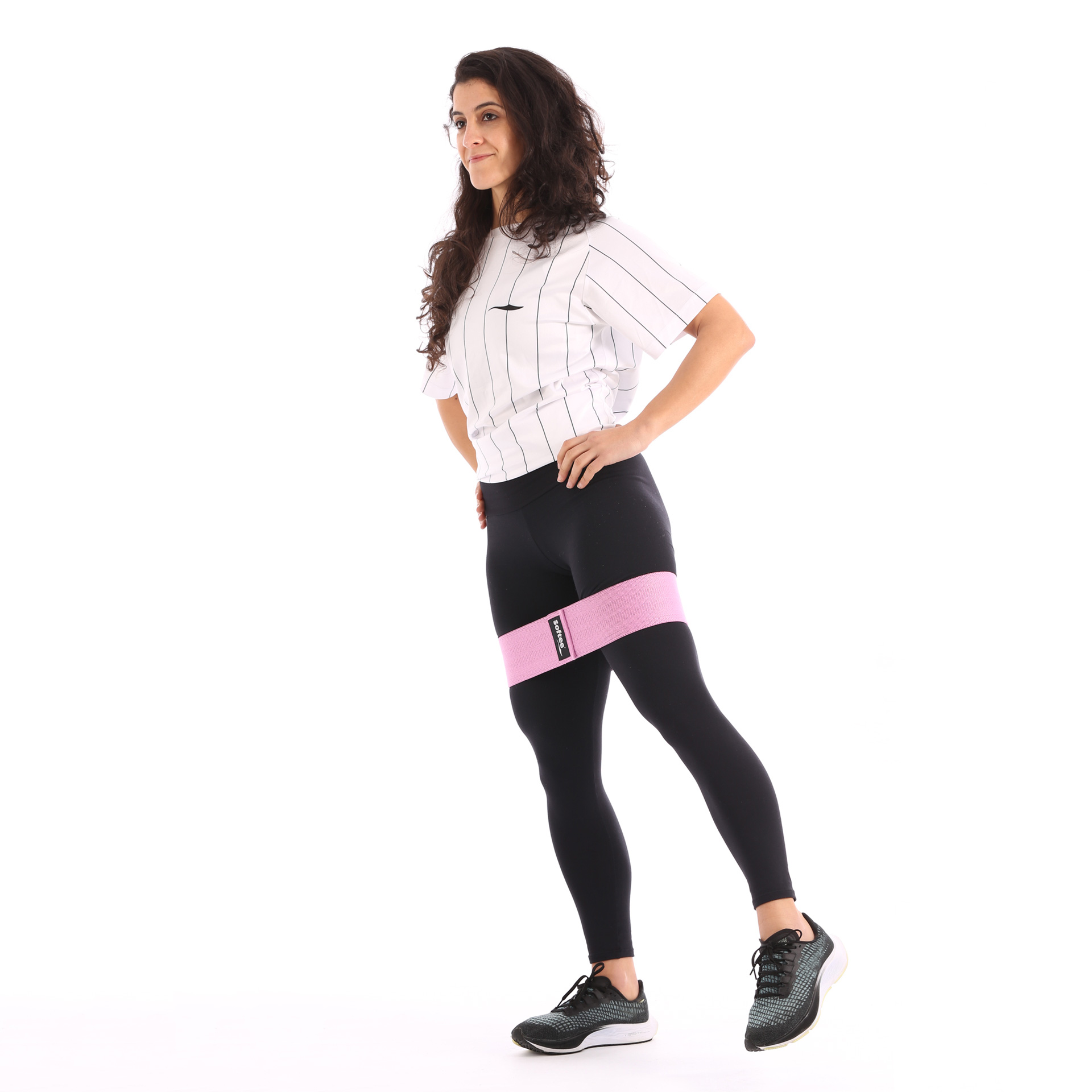 Mujer en ropa deportiva con banda de resistencia en los muslos. Está de pie sobre un fondo blanco.
