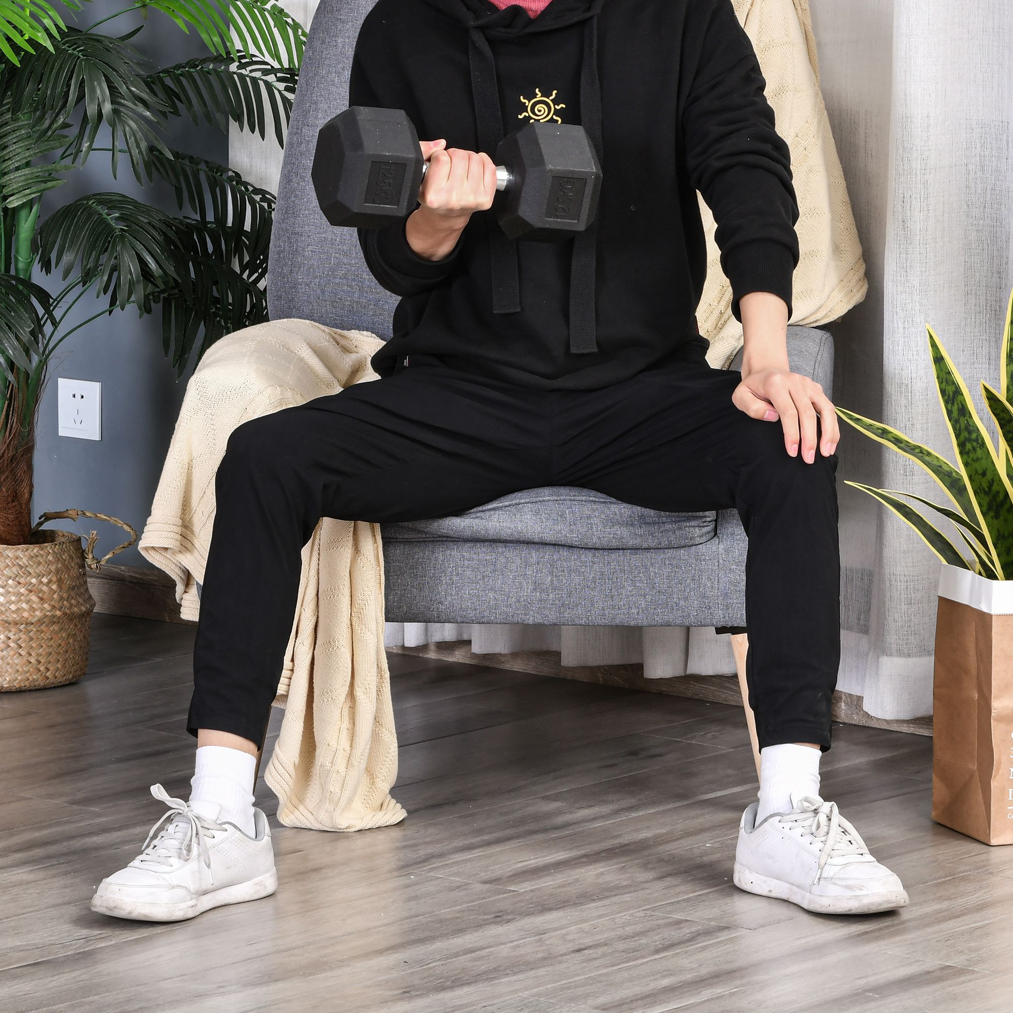 Persona con chándal negro levanta una mancuerna de 12,5 kg mientras está sentada en una silla gris. Plantas, manta y zapatillas blancas son visibles.