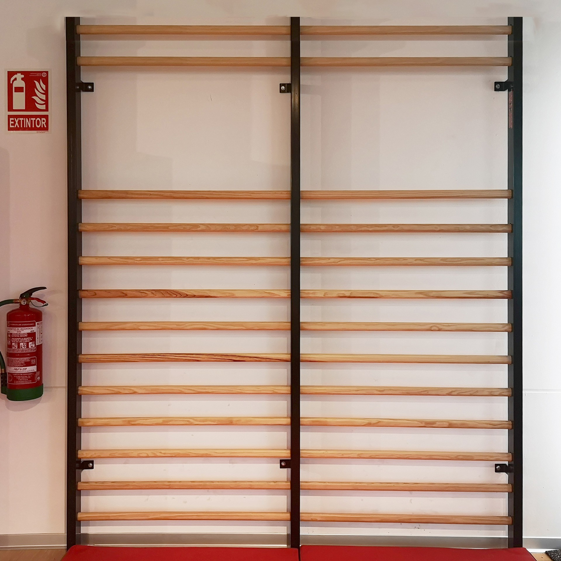 Una scala in legno montata a parete con supporti metallici. Un estintore e un cartello a sinistra. Un tappeto rosso in basso.