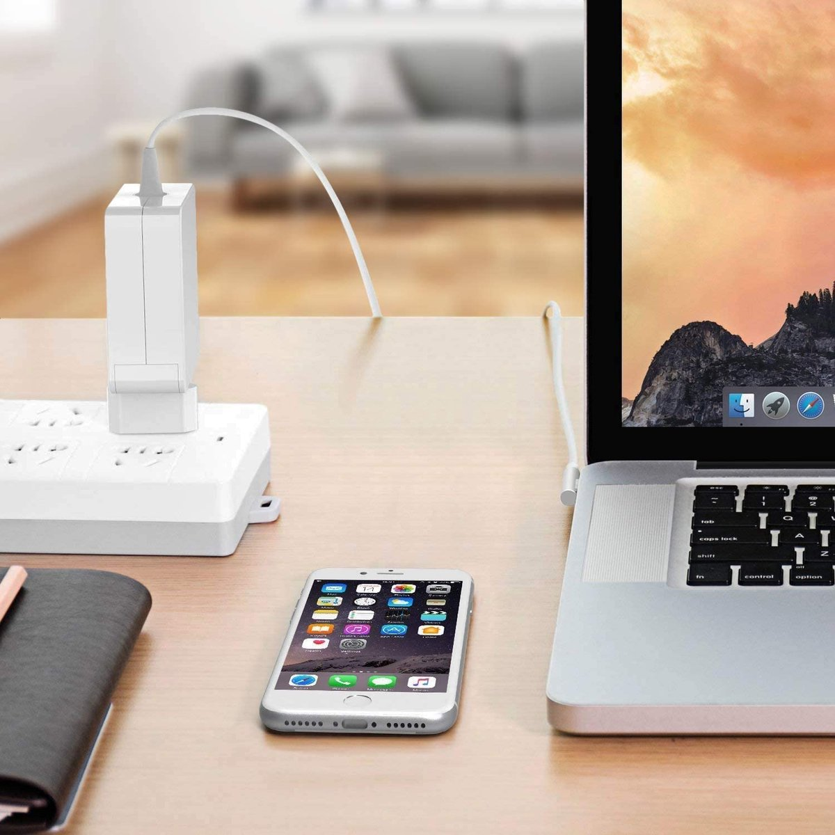 A laptop, phone, and charger on a wooden desk. The phone is in the foreground, laptop in the background. A white charger is plugged in.