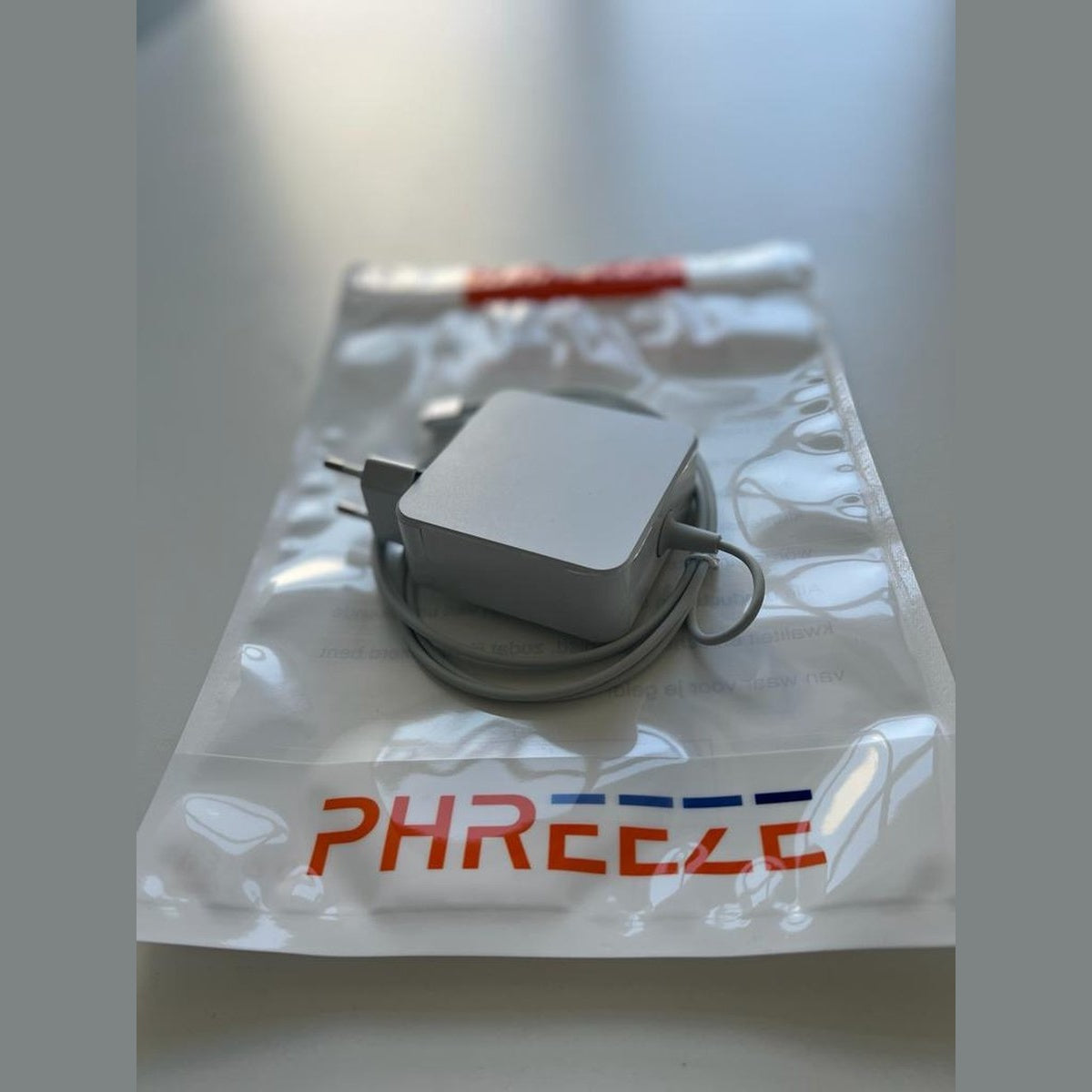 Een witte oplader met een grijze kabel zit in een doorzichtige plastic zak. De zak heeft het woord 'PHREEZE' in rood en blauw.