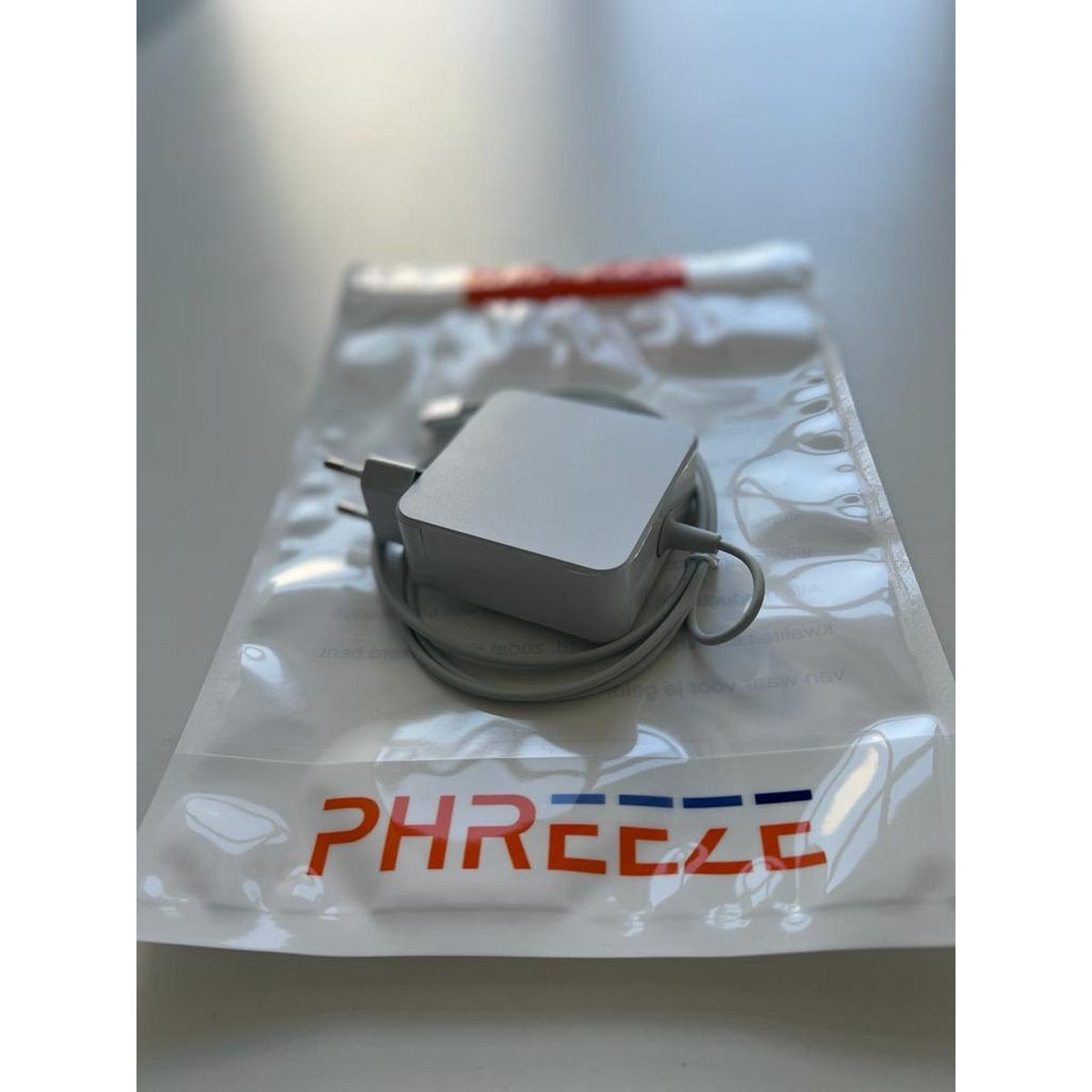 Een witte voedingsadapter met een grijze kabel, in een doorzichtige plastic zak. De zak heeft rode en blauwe tekst: PHREEZE.