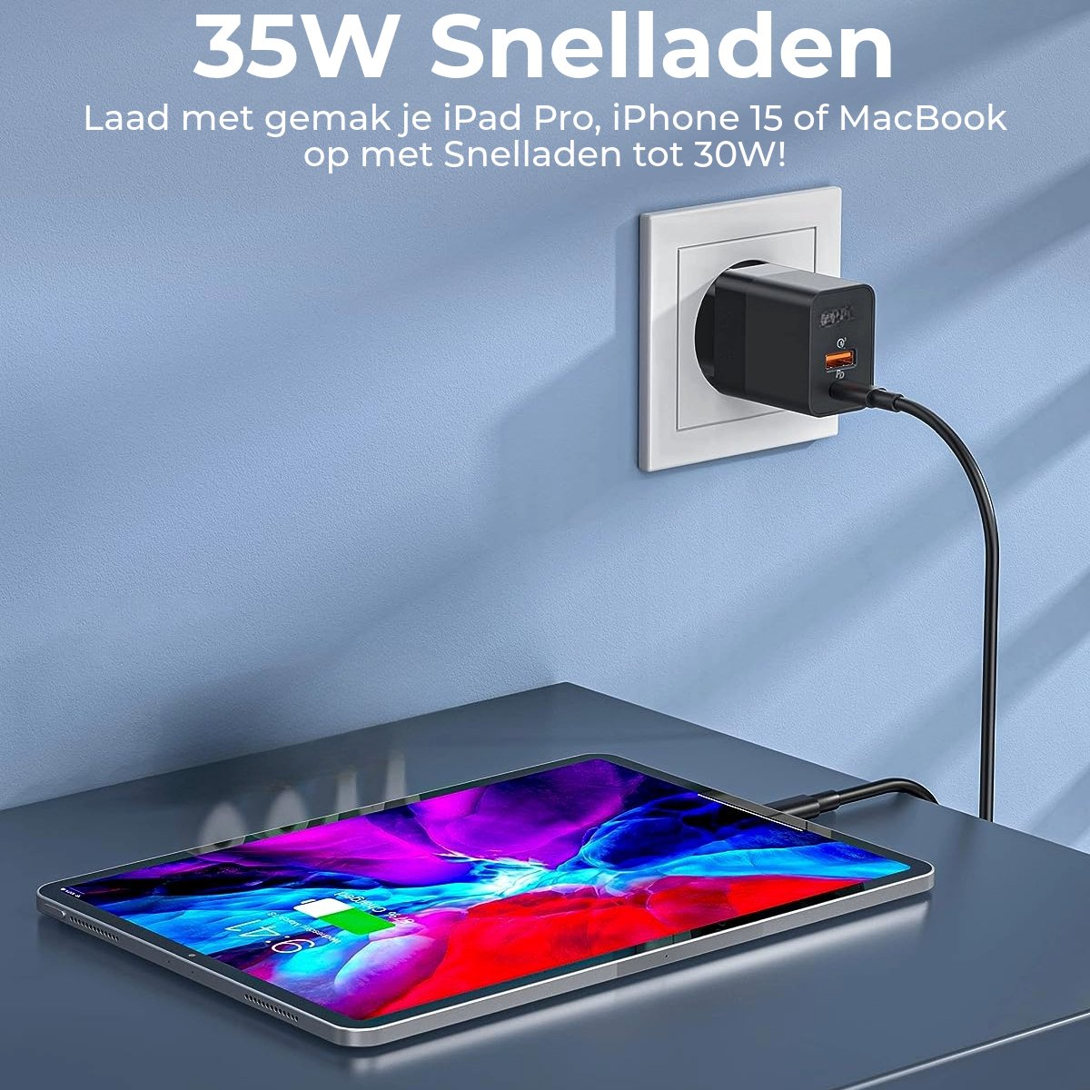 An iPad Pro charging with a black charger in a wall outlet. The tablet is on a table with a blue wall in the background.