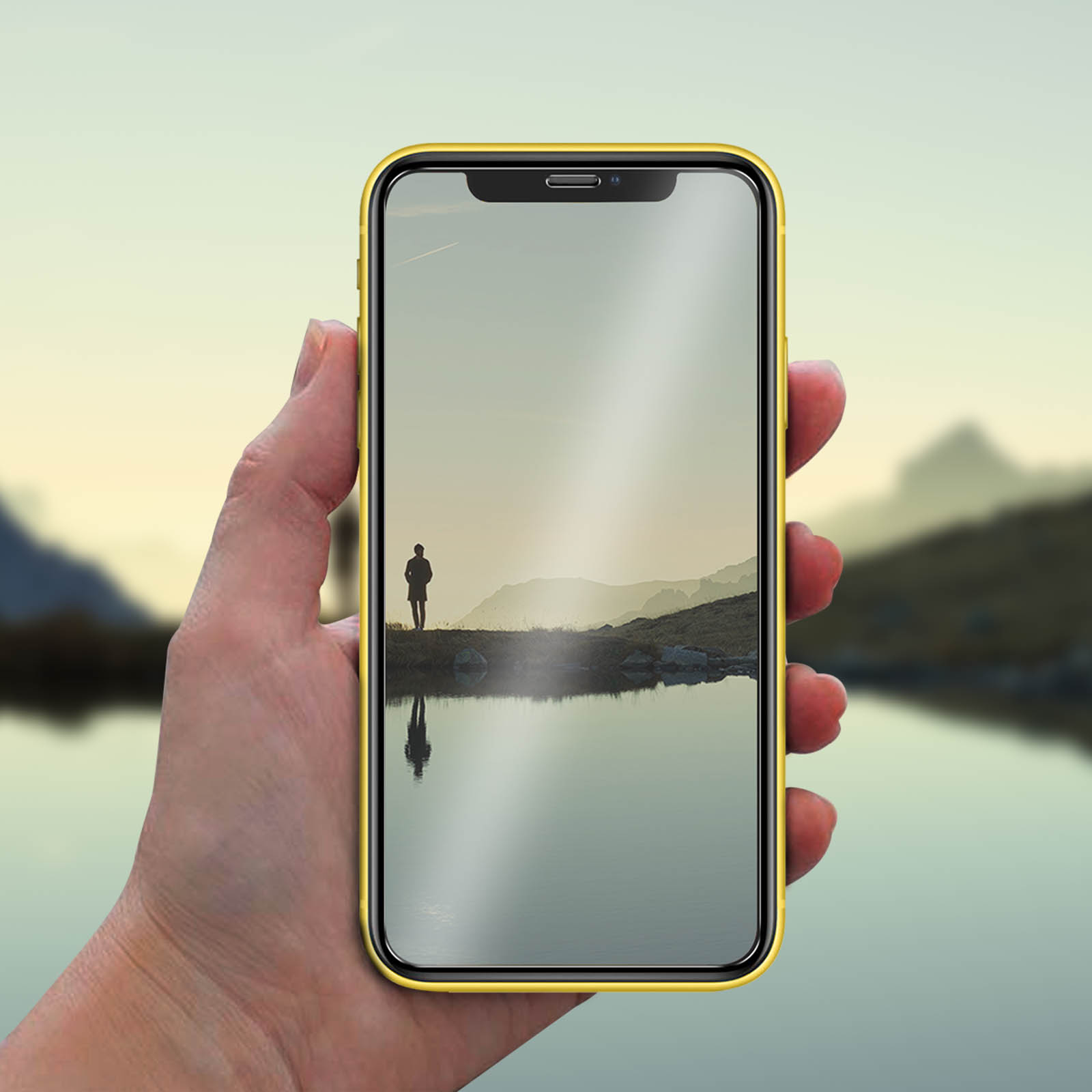 Mano che tiene un telefono con vista sul paesaggio. Una persona è in piedi su una riva che si riflette nell'acqua. Custodia gialla.