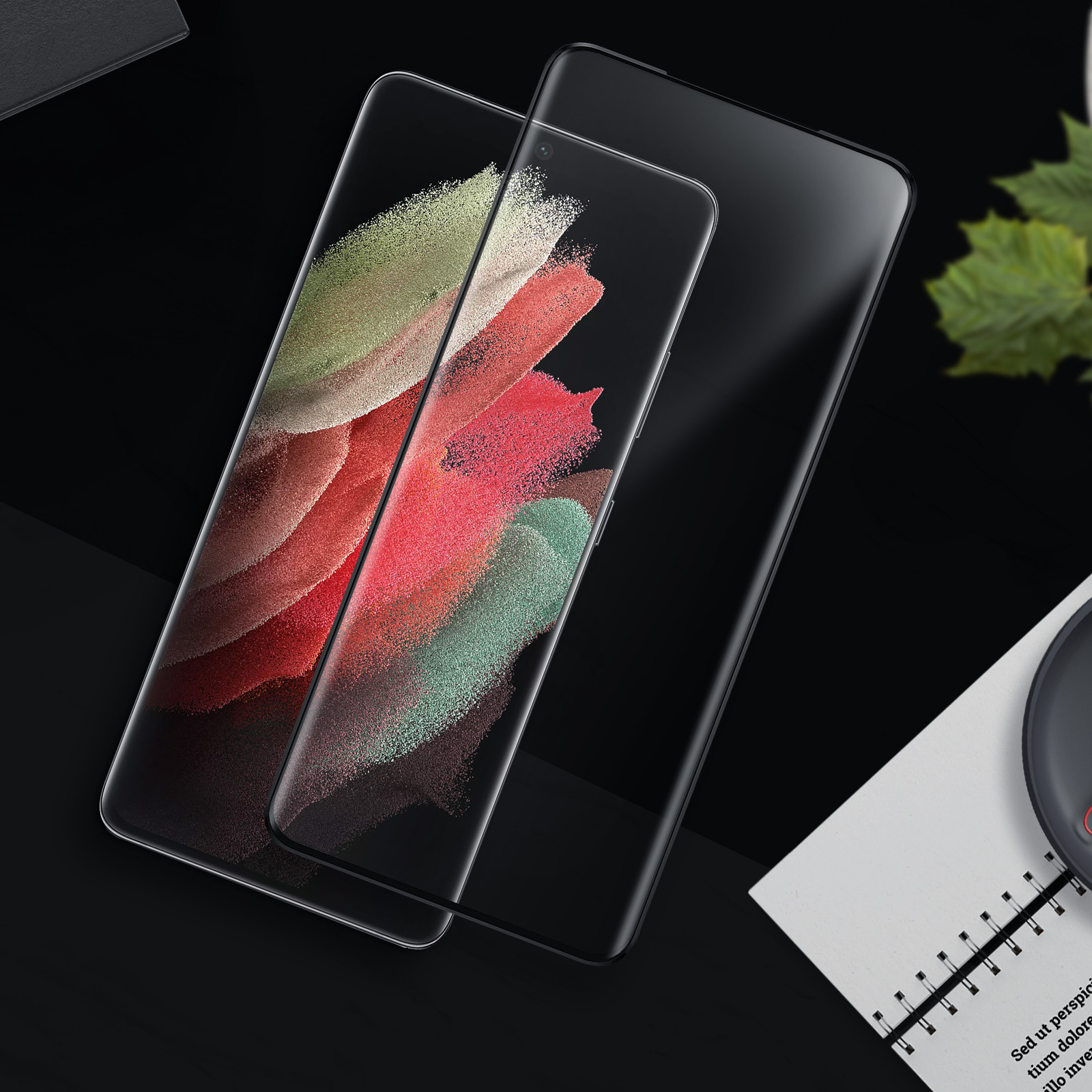 Due smartphone su superficie nera. Uno mostra arte astratta colorata, l'altro nero. Taccuino e pianta sullo sfondo.