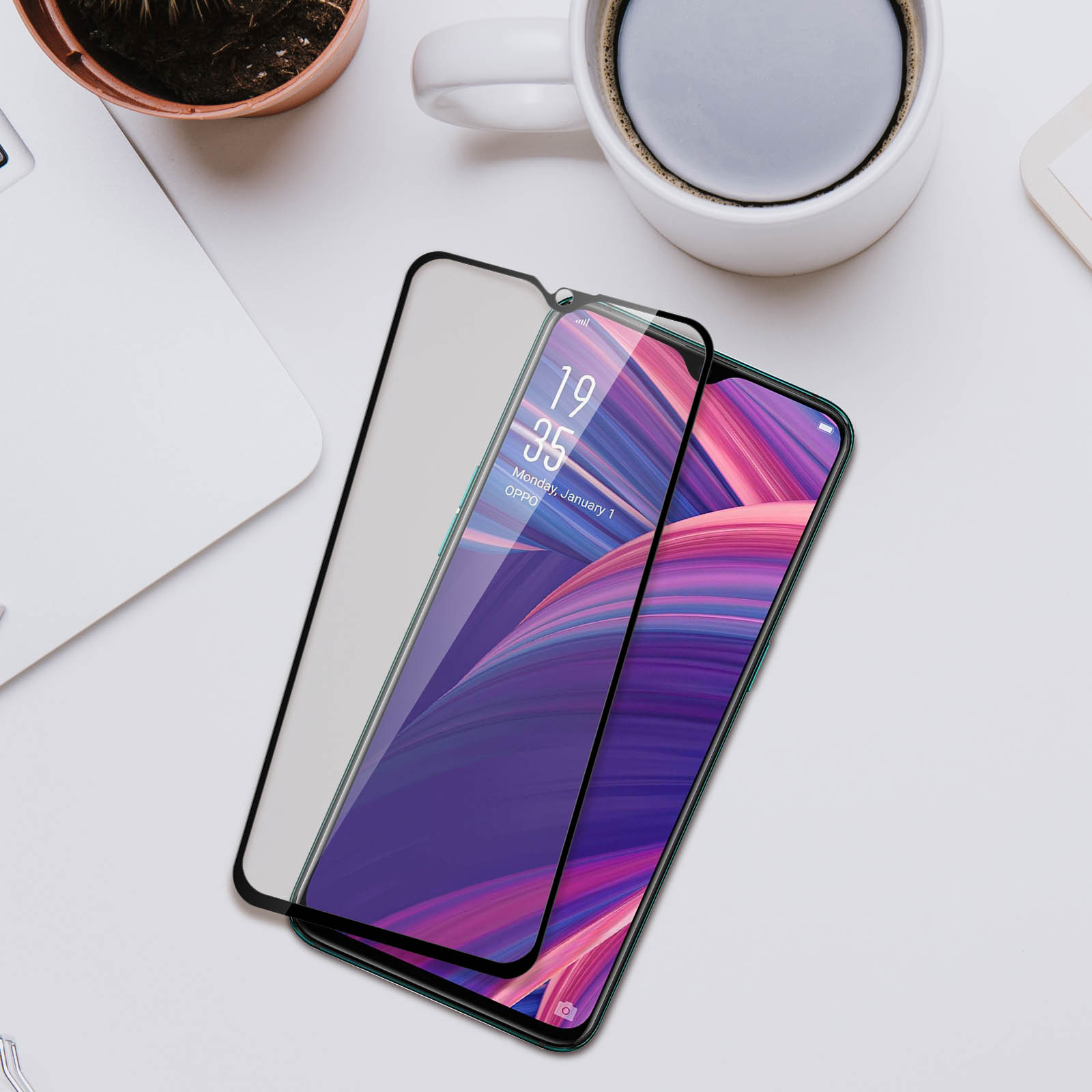 Uno smartphone con protezione schermo su una superficie bianca, con tazza di caffè, laptop e pianta sullo sfondo.