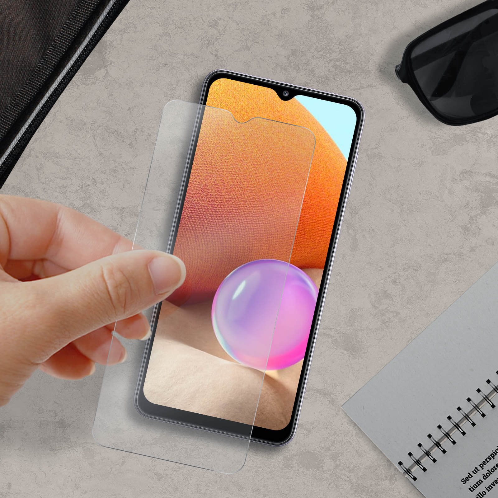 Una mano tiene una protezione per schermo sopra uno smartphone. Occhiali da sole, un taccuino e una borsa nera sono sullo sfondo.