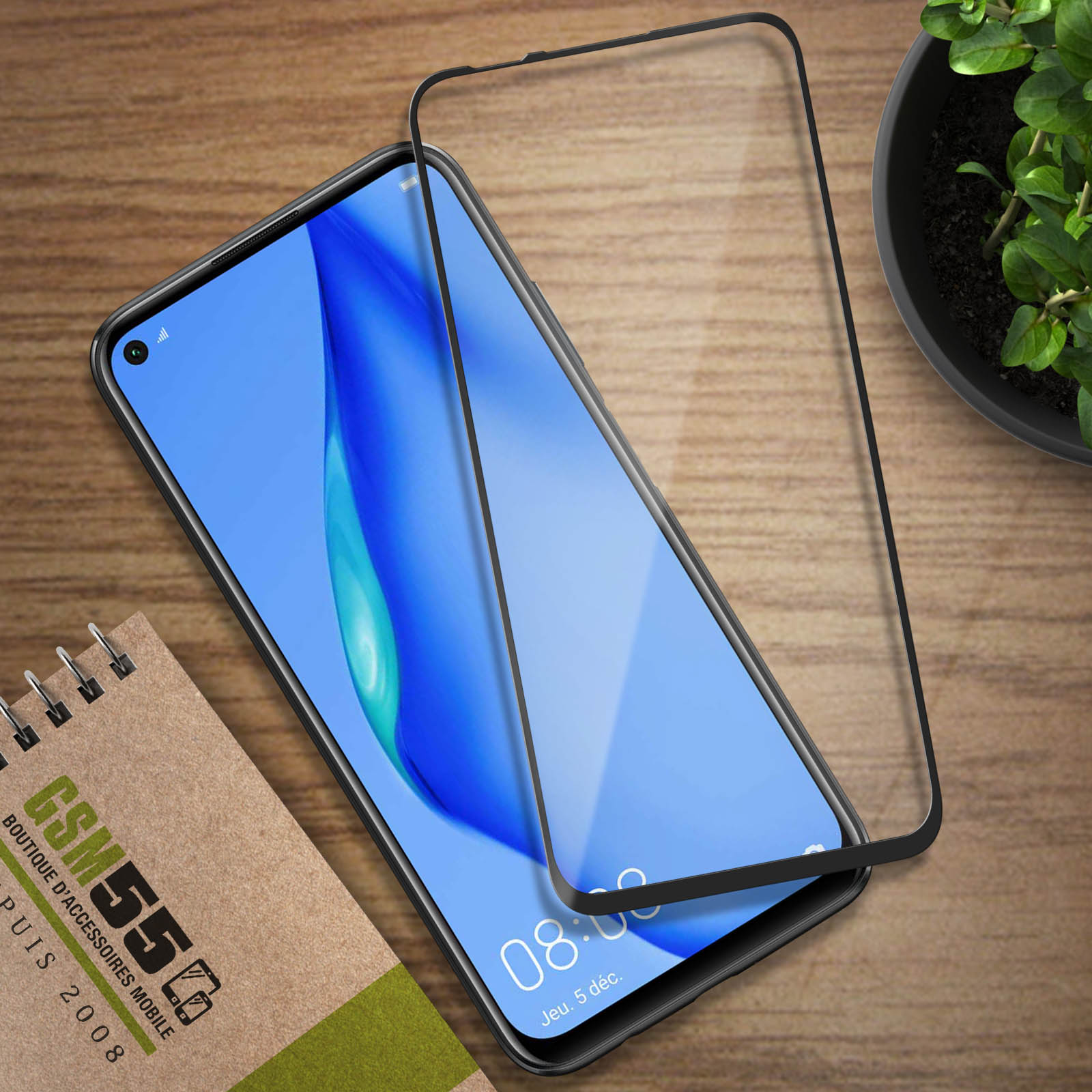 Uno smartphone con protezione schermo, blocco note e una pianta in vaso su una superficie di legno.