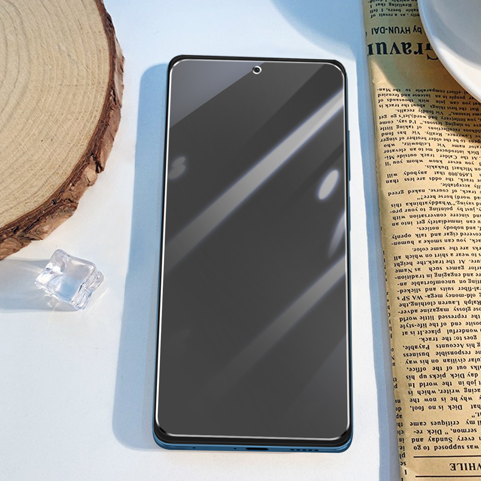 Un proteggi schermo per smartphone su un giornale con un tagliere e un cubetto di ghiaccio accanto.