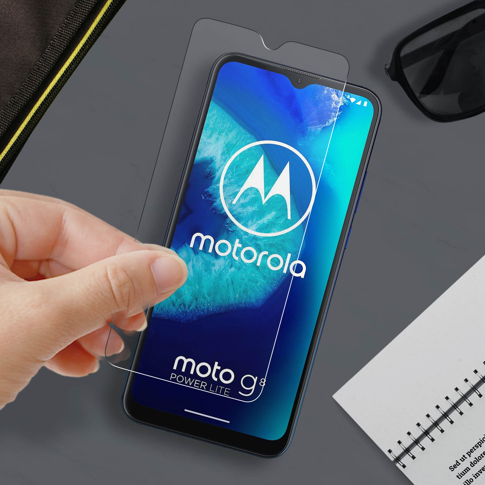 Una mano applica una protezione per lo schermo a un telefono Motorola Moto g8 Power Lite. Sono visibili anche occhiali da sole e un quaderno.