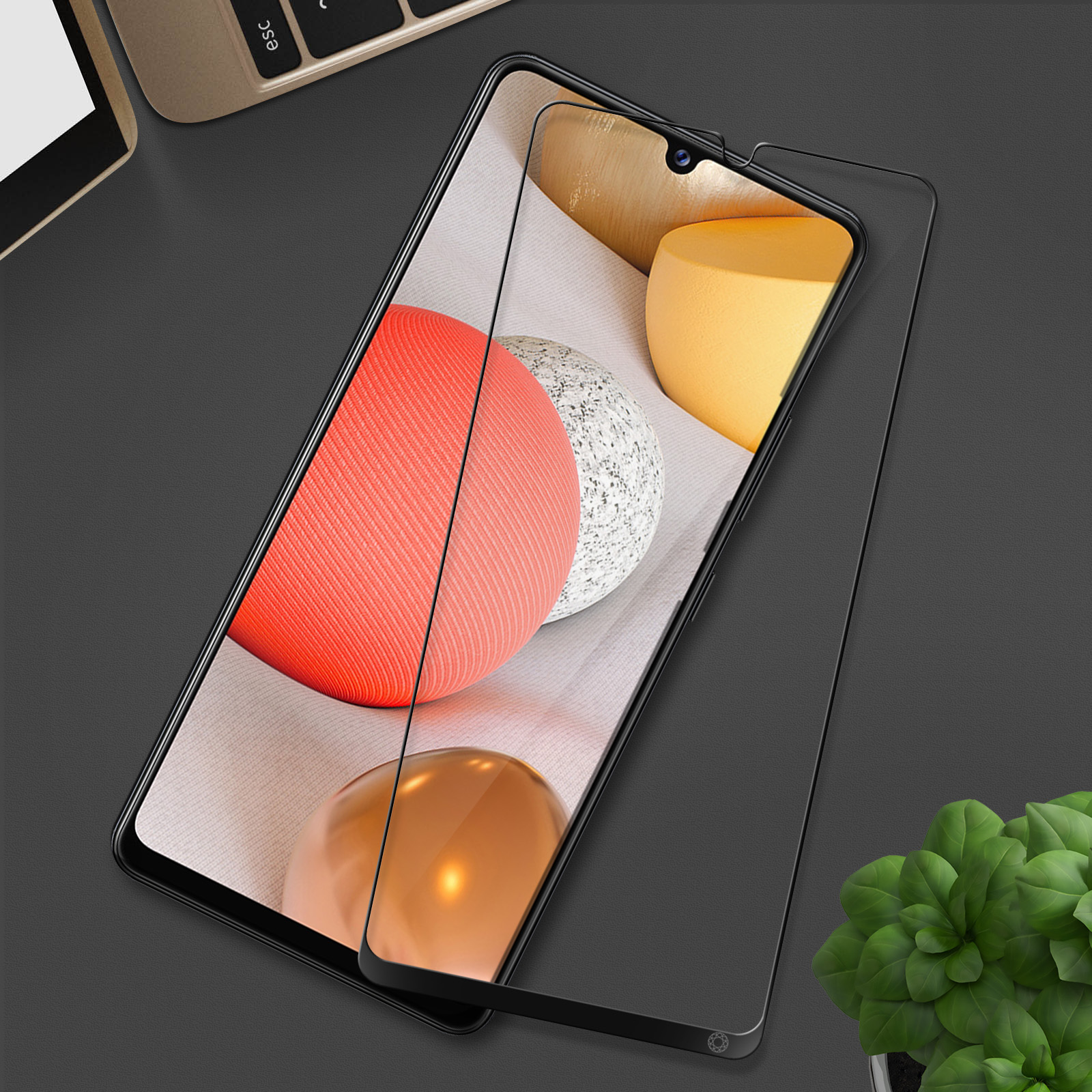 Un écran de téléphone avec un protecteur. L'écran affiche des sphères rouges, blanches et jaunes. Un ordinateur portable et une plante en arrière-plan.