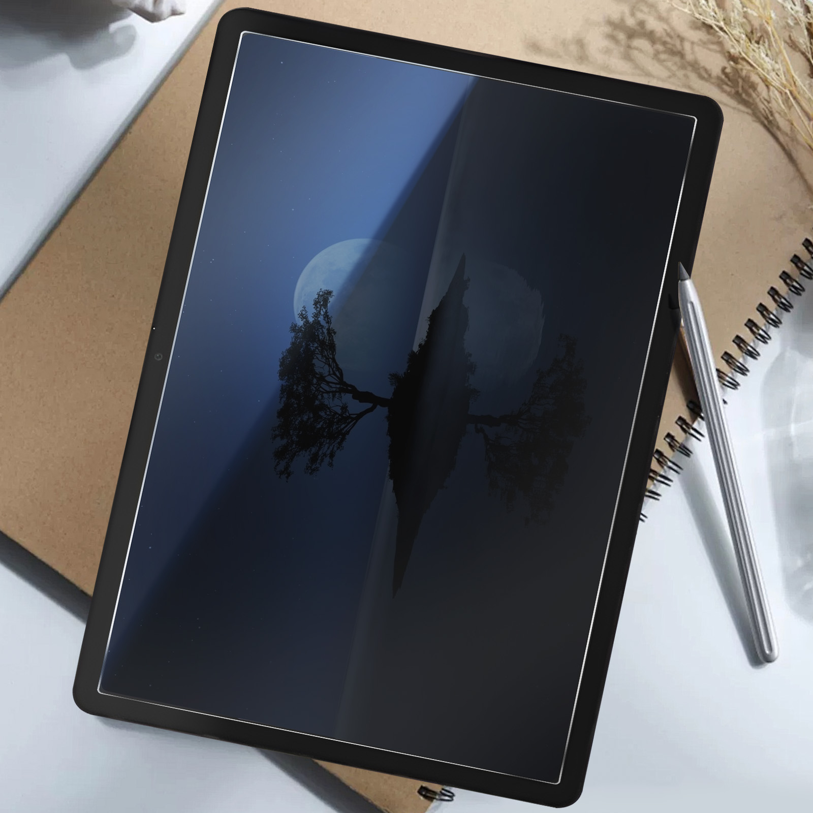 Un tablet mostra la sagoma di un albero contro un cielo illuminato dalla luna. È appoggiato su un quaderno, con una penna e piante secche vicine.