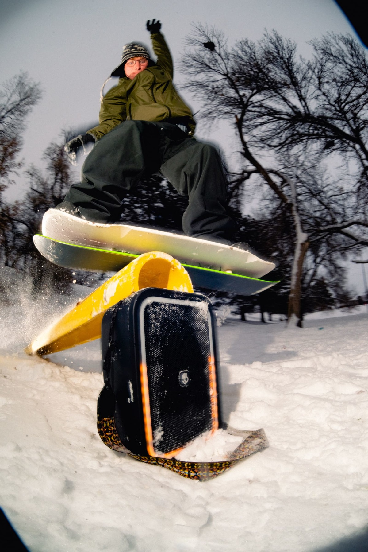 Snowboarder in der Luft über einem schwarzen Koffer und einer gelben Schiene. Schnee wird aufgewirbelt. Kahle Bäume sind im Hintergrund.